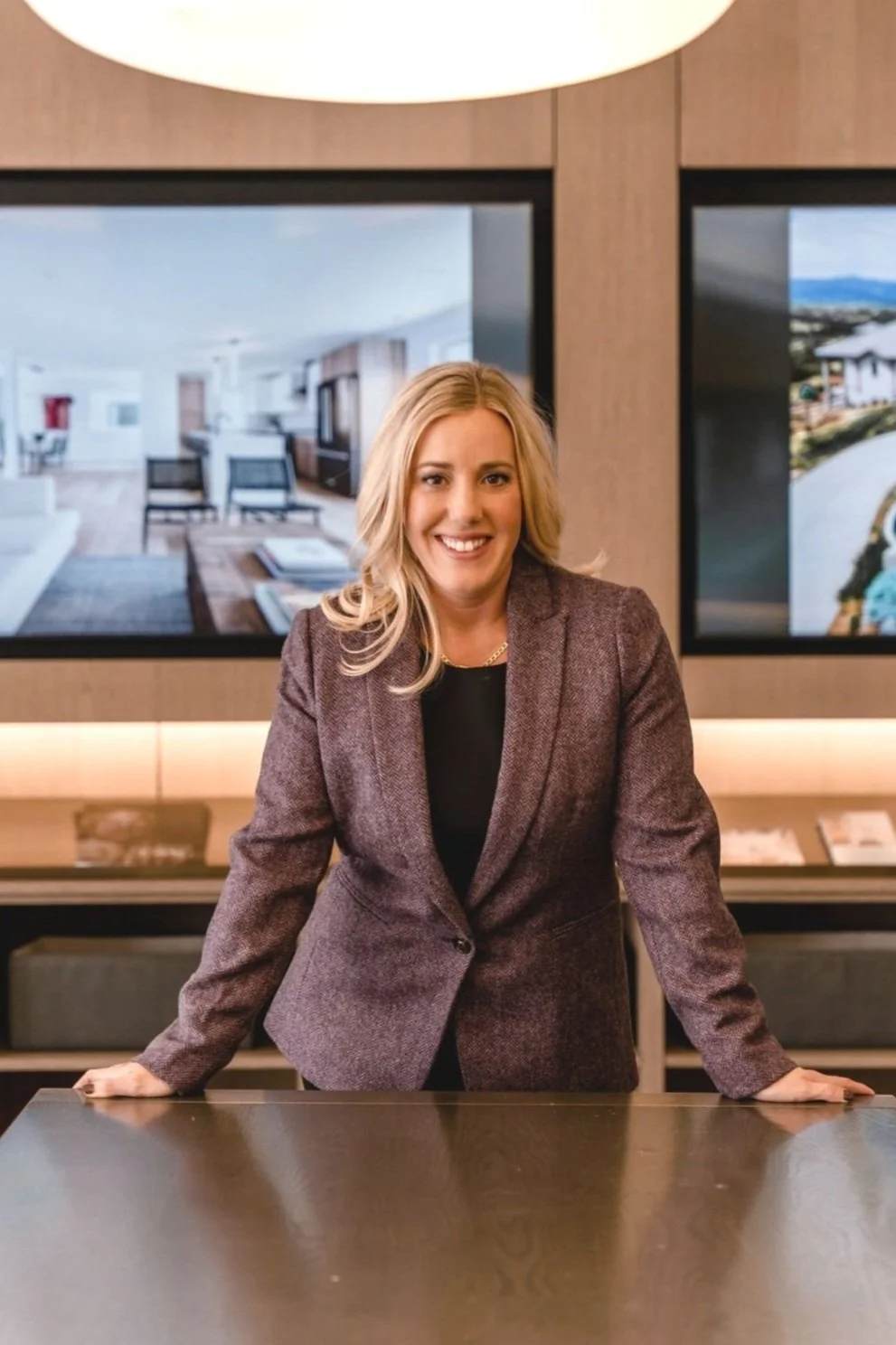 A smiling woman in a brown blazer standing behind a dark table in a modern office or conference room with two large screens behind her showing interior and exterior images.