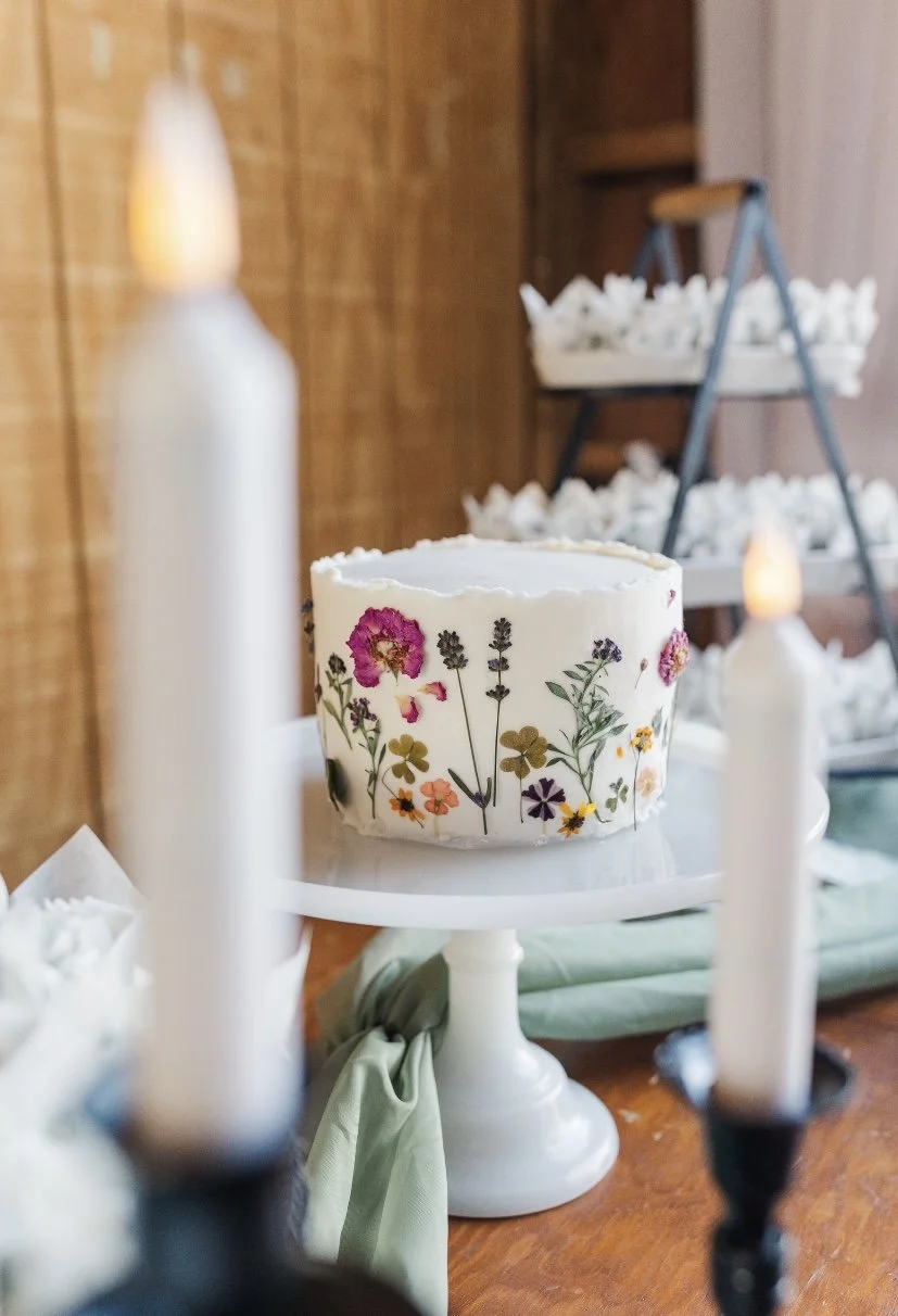 A round white cake with floral decorations on the side, displayed on a white cake stand in a warmly lit room with candles and wooden furniture.