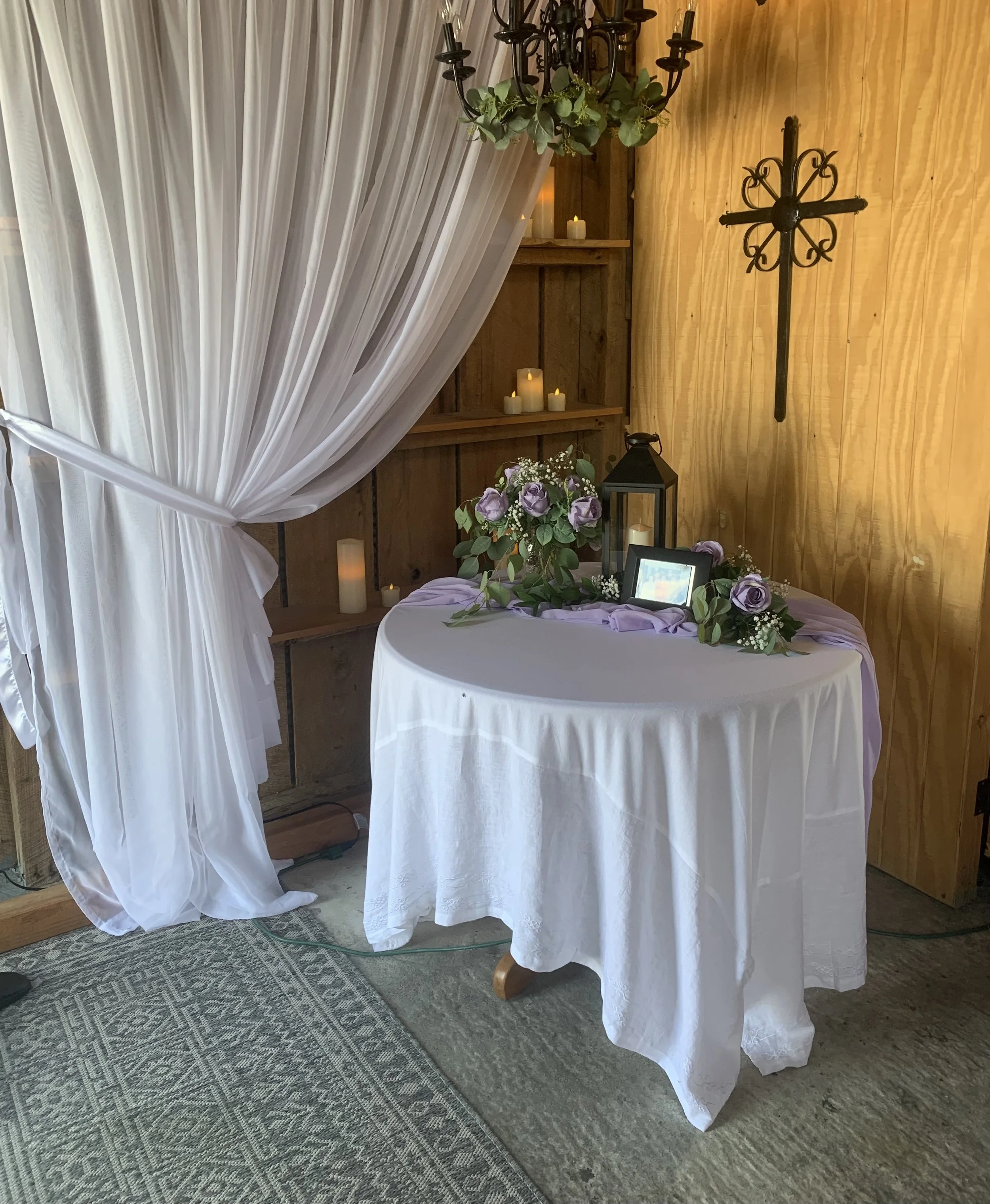 A table with a white tablecloth, decorated with a floral arrangement of purple roses, candles, a lantern, and a framed photo, set against a wooden wall with candles and a cross.
