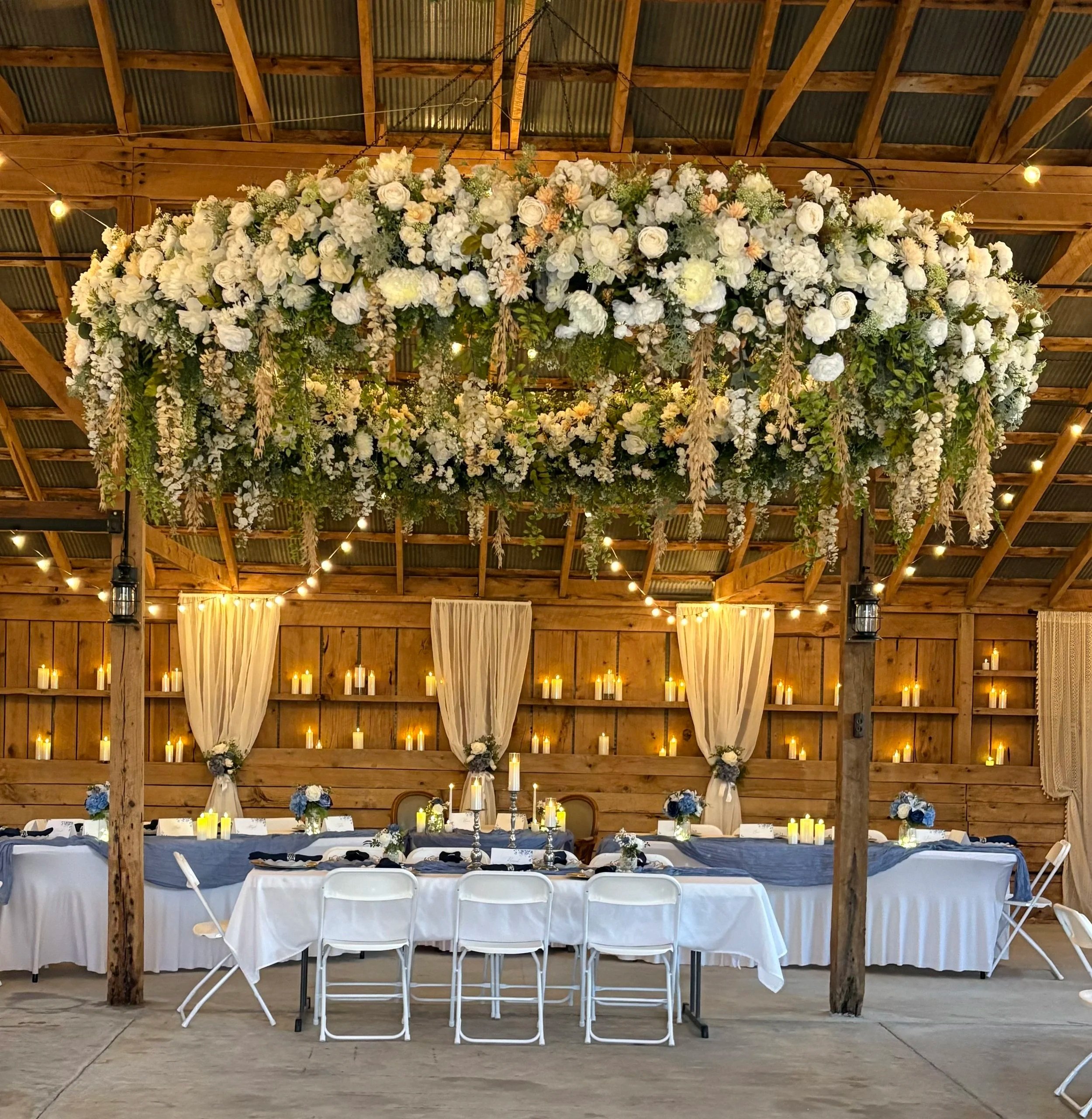 Decorated wedding reception with a large floral chandelier, candles, and draped curtains in a rustic barn setting.