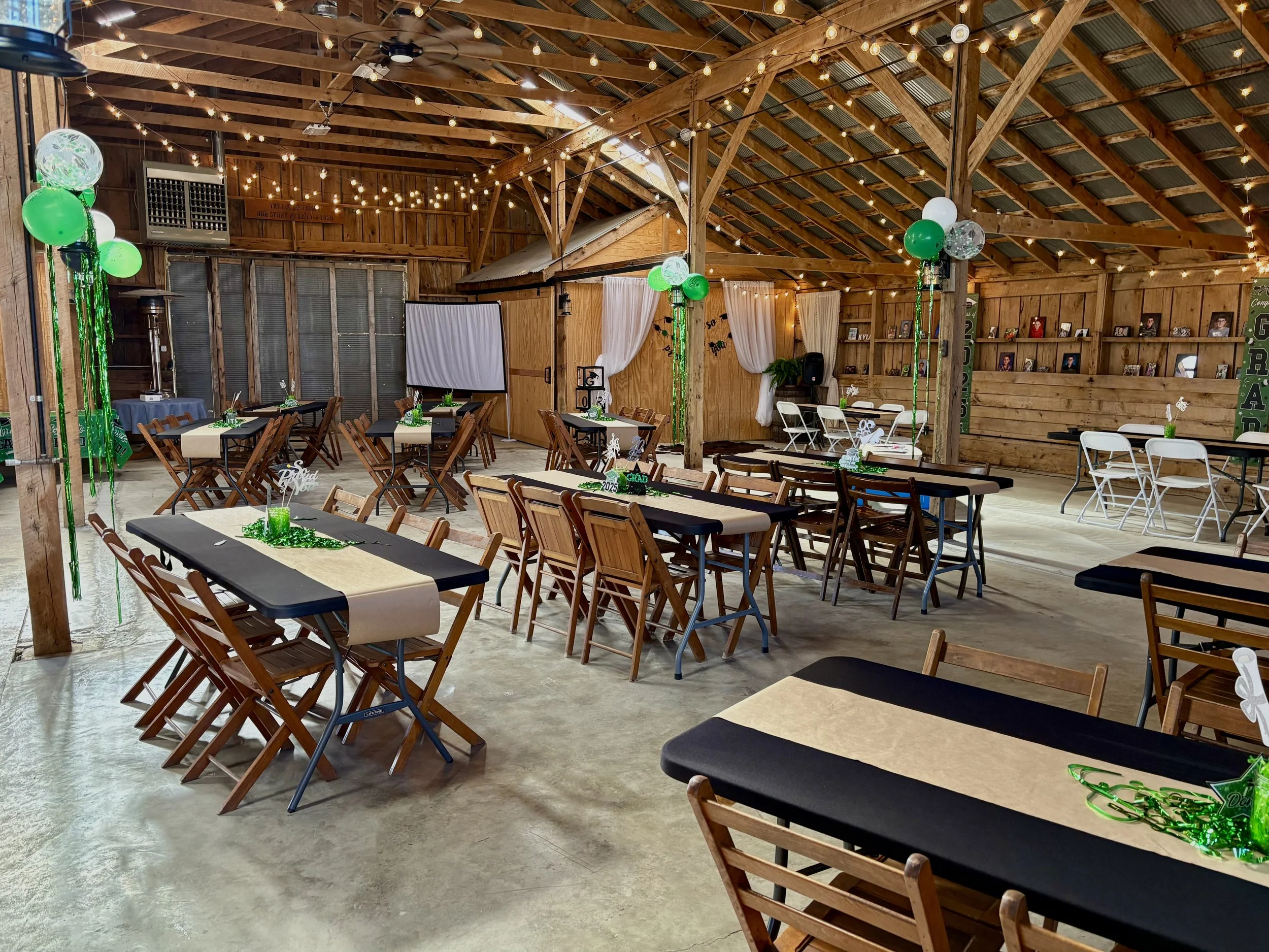 Decorated rustic barn interior with tables and chairs for a celebration, green and white balloons, string lights, and festive decorations.