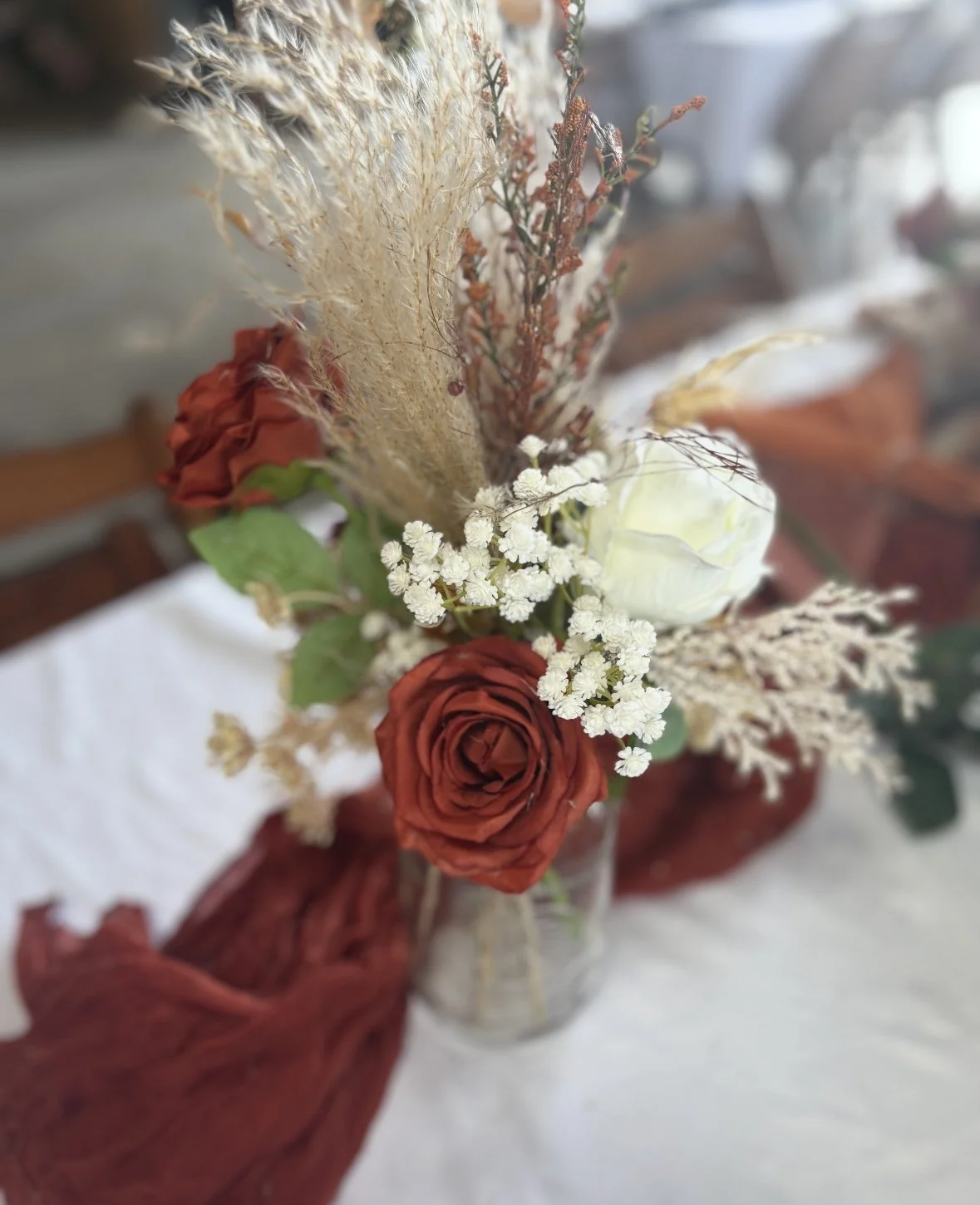 A floral arrangement featuring red roses, white flowers, dried pampas grass, and brown and green foliage in a glass vase on a white tablecloth.