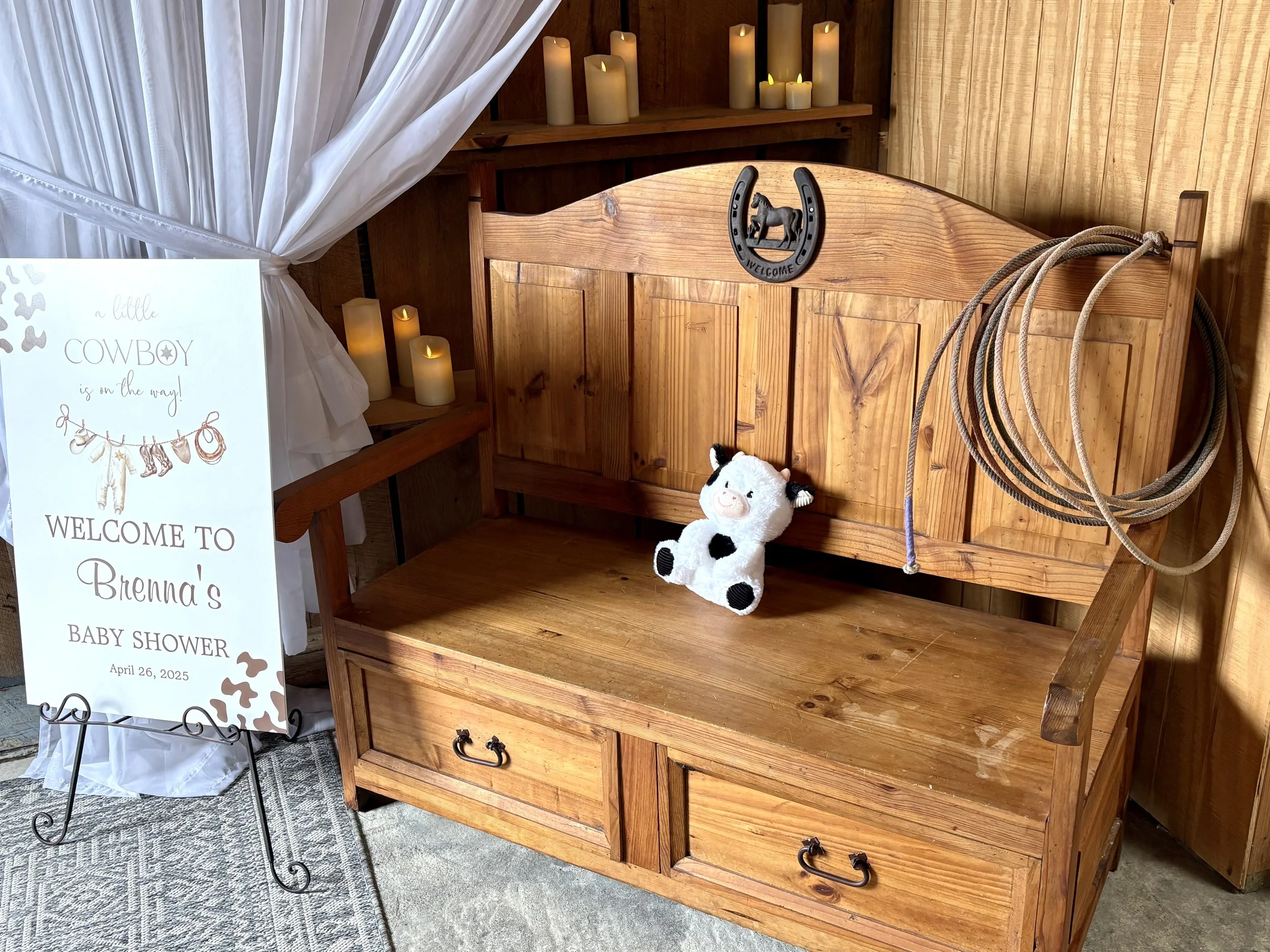 A wooden bench with a plush cow toy sitting on it, and a black horseshoe decoration above it. To the right, a bundle of horse riding lassos. To the left, a sign welcoming guests to Brenna's baby shower with a cowboy theme, decorated with a cow and ch