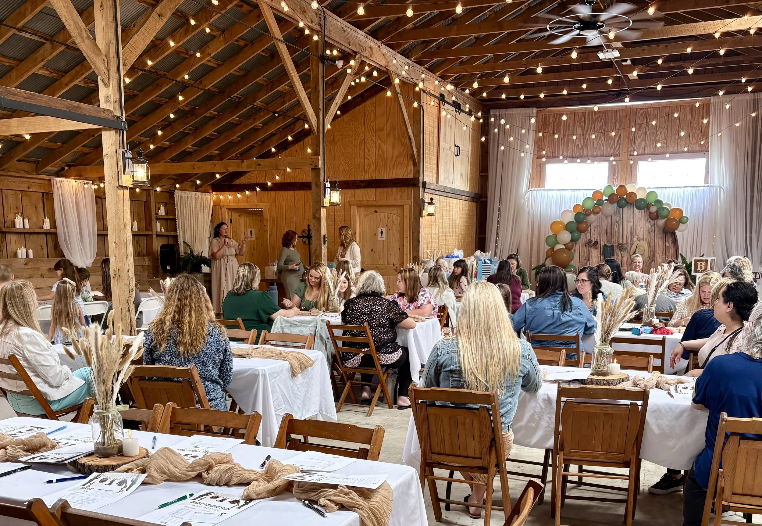 Women gathered in a rustic barn decorated for an event with string lights, balloons, and table settings for a celebration or meeting.