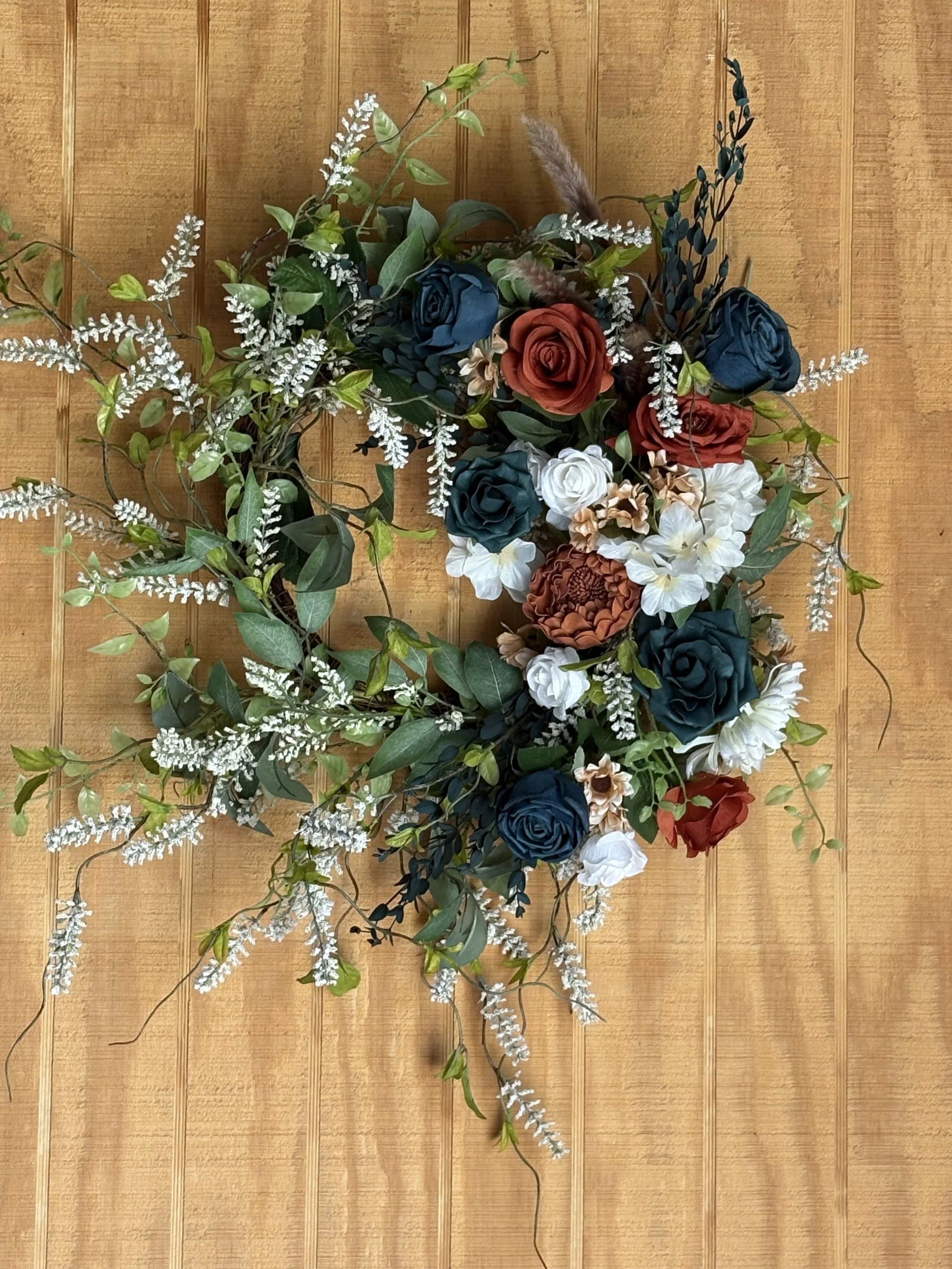 A floral wall hanging with blue, white, red, and orange flowers, greenery, and white accent flowers on a wooden paneled background.