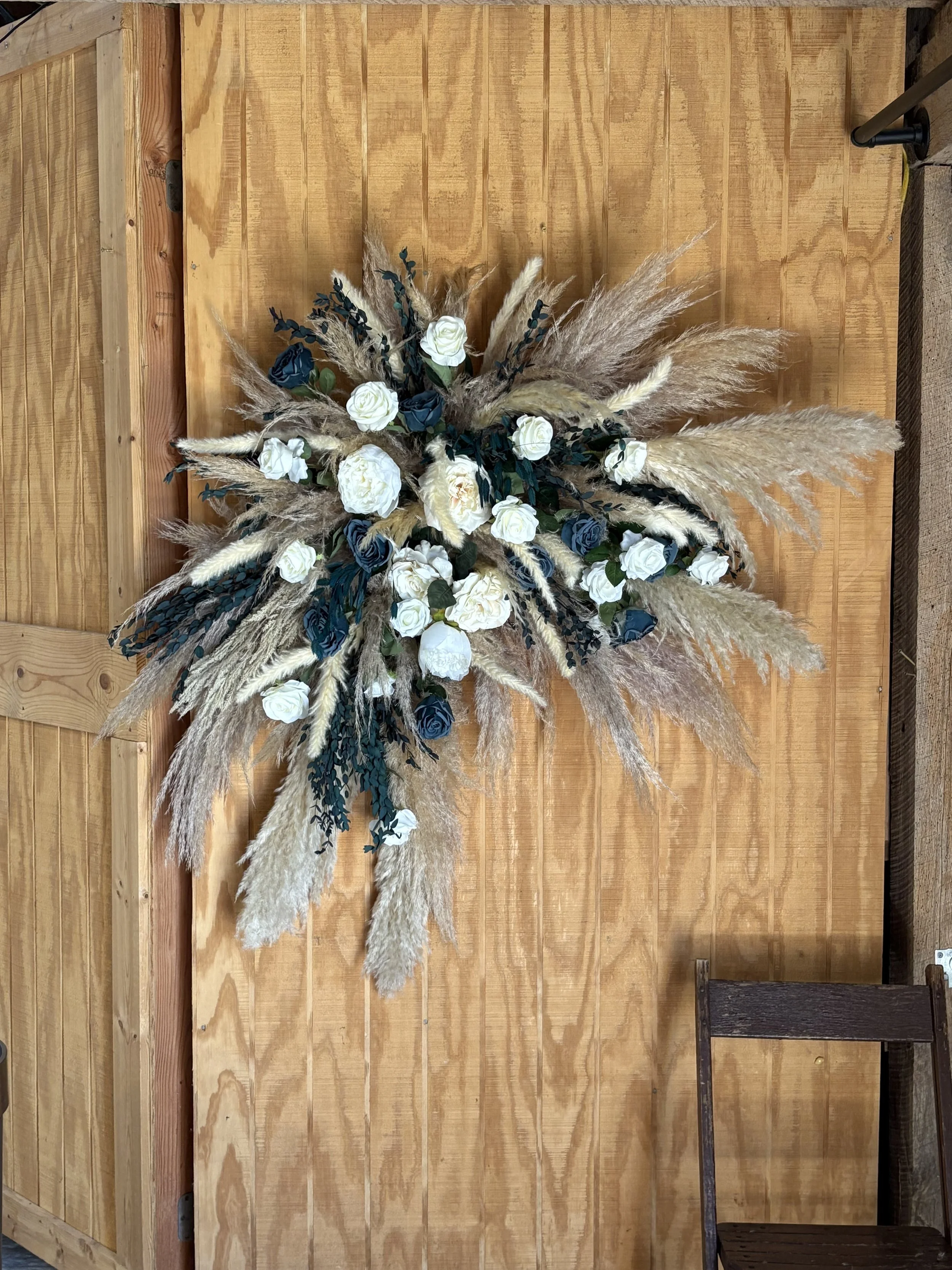A large floral arrangement with white roses, dark blue roses, and pampas grass on a wooden wall.