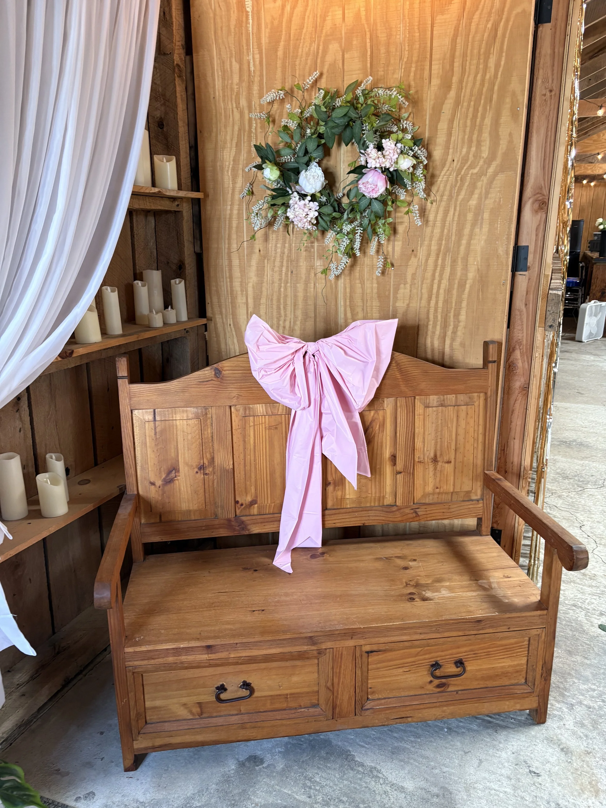 Wooden bench with a pink bow on the backrest, placed in front of a wooden wall decorated with a floral wreath. Candles on shelves are on the side.