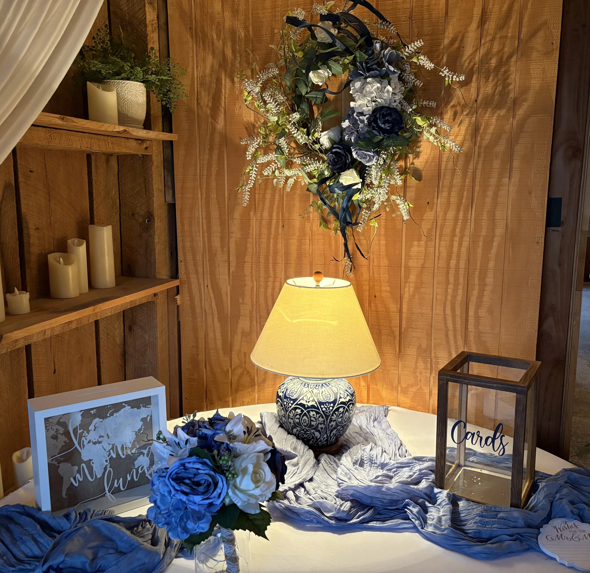 A decorative table setup including a floral arrangement, a table lamp, a framed world map with the words 'where we will fun,' a bouquet of blue and white flowers, a card box labeled 'Cards,' and a wooden shelf with candles against a wood-paneled wall