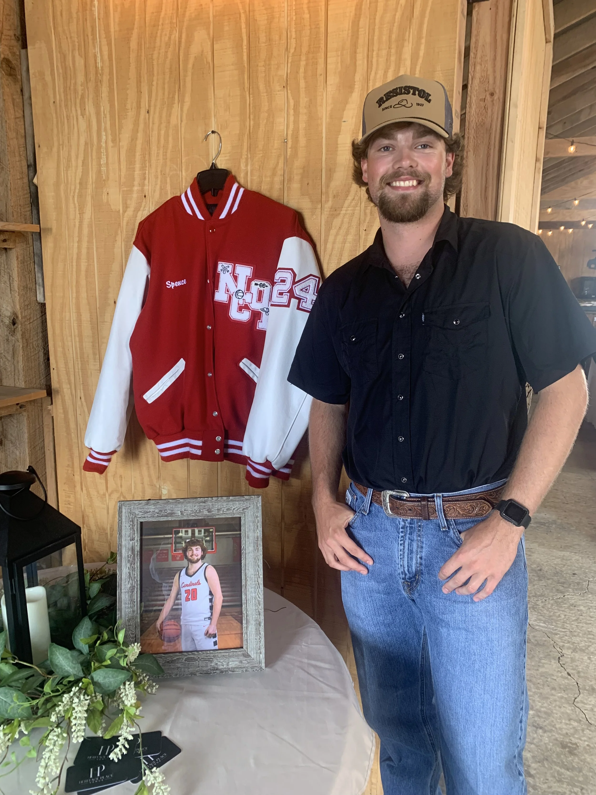 A man standing next to a red and white letterman jacket and a framed photo of a basketball player. The man is smiling, wearing a black shirt, blue jeans, a beige cap, and a watch. The photo is in a rustic setting with wooden walls.