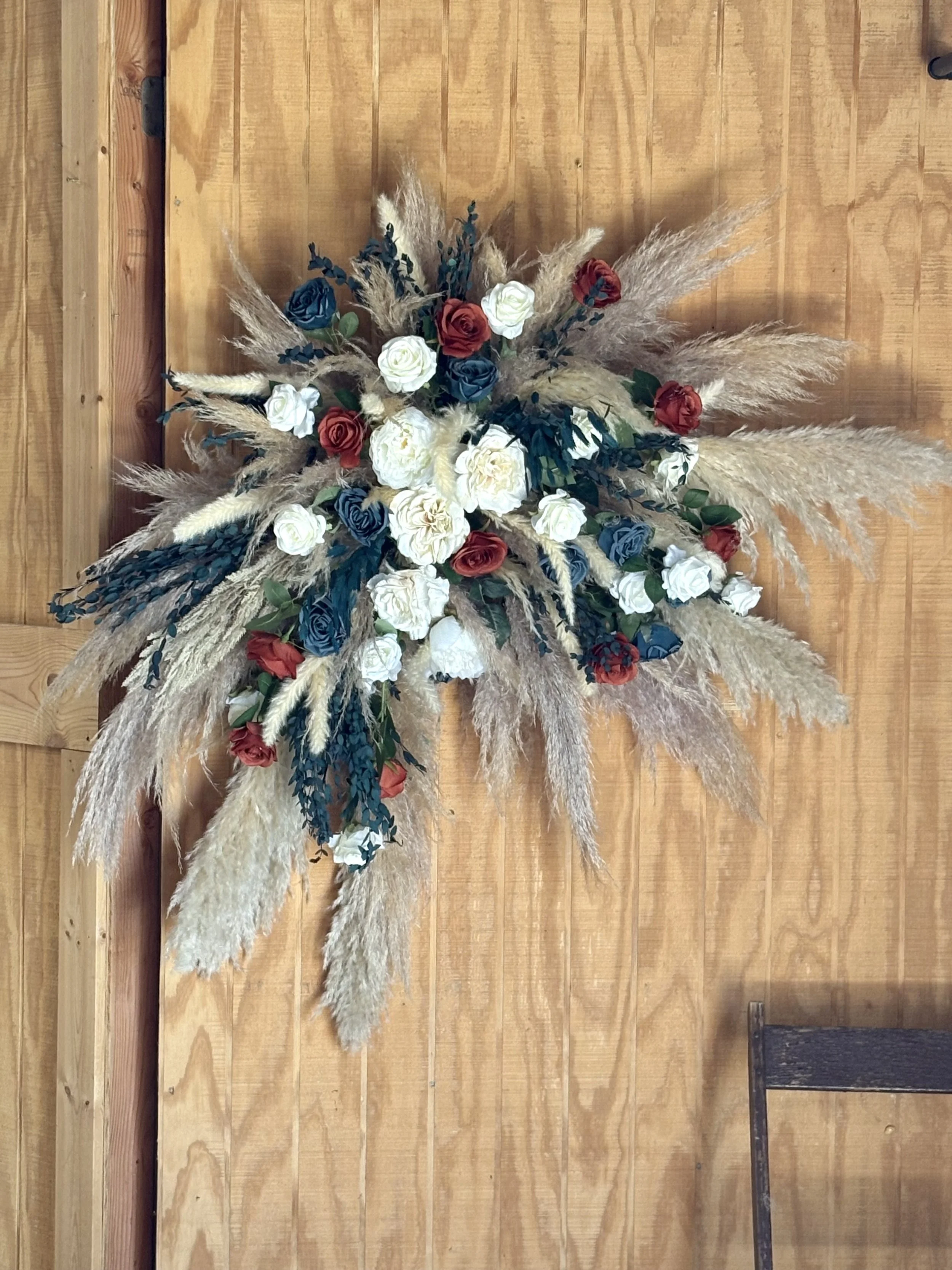 A decorative floral arrangement with white, red, and blue roses, alongside beige pampas grass and greenery, mounted on a wooden wall.