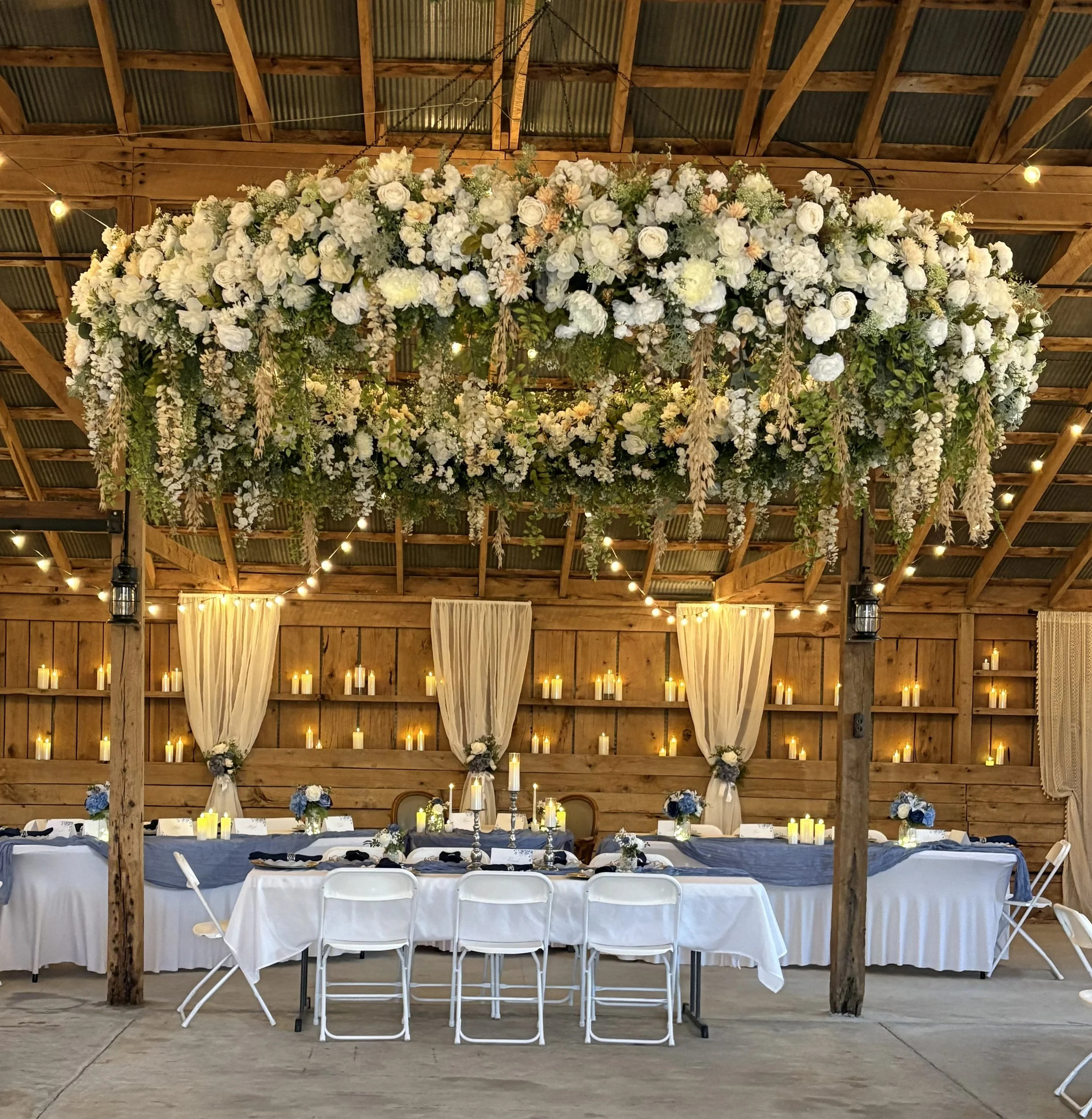 Elegant wedding reception setup in a rustic barn with a long head table decorated with blue and white flowers, surrounded by white chairs, with a large floral chandelier hanging overhead, candlelit backdrop, and soft string lights.