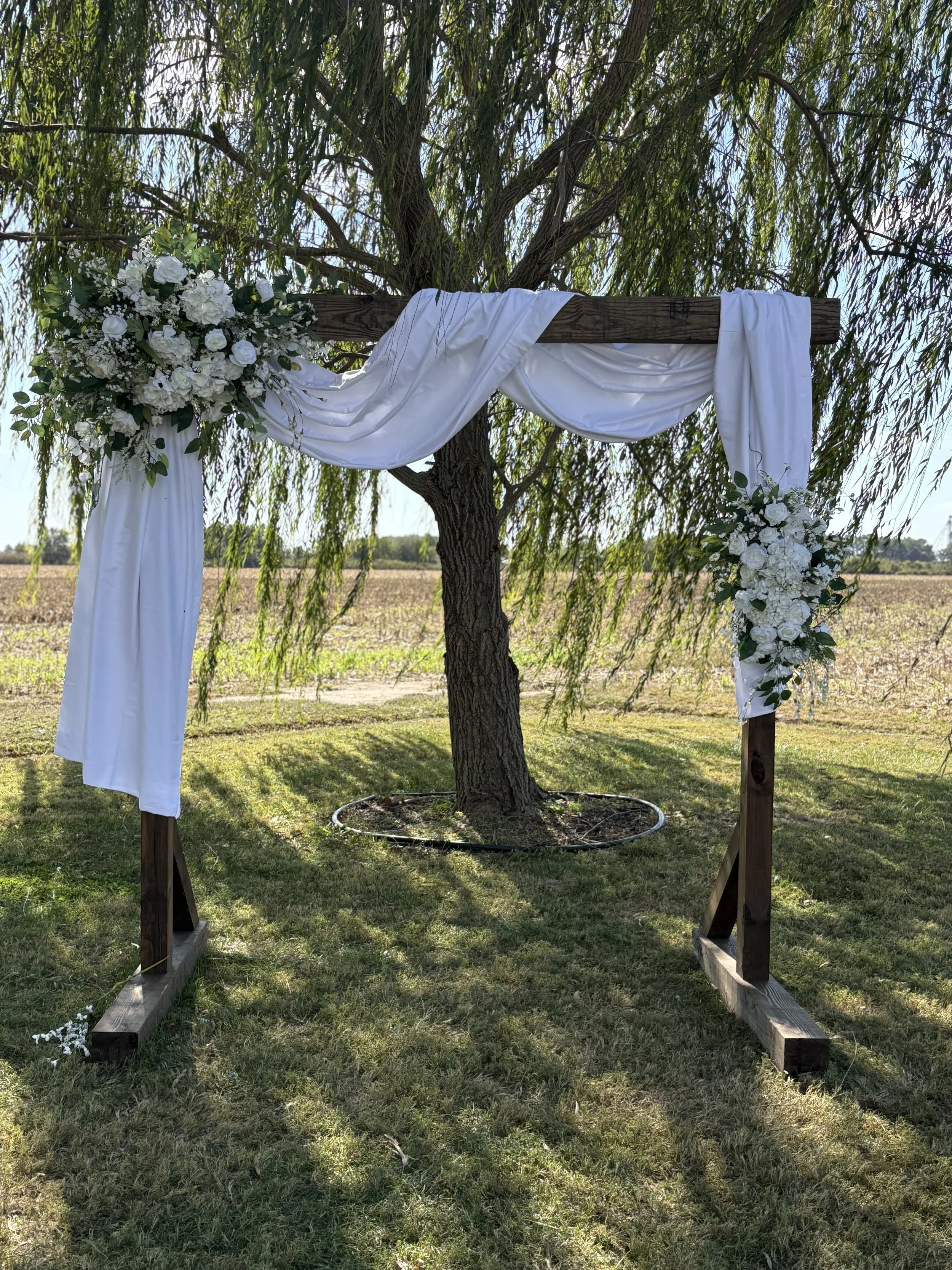 A wedding arch decorated with white drapes and floral arrangements stands under a tree in an outdoor setting.