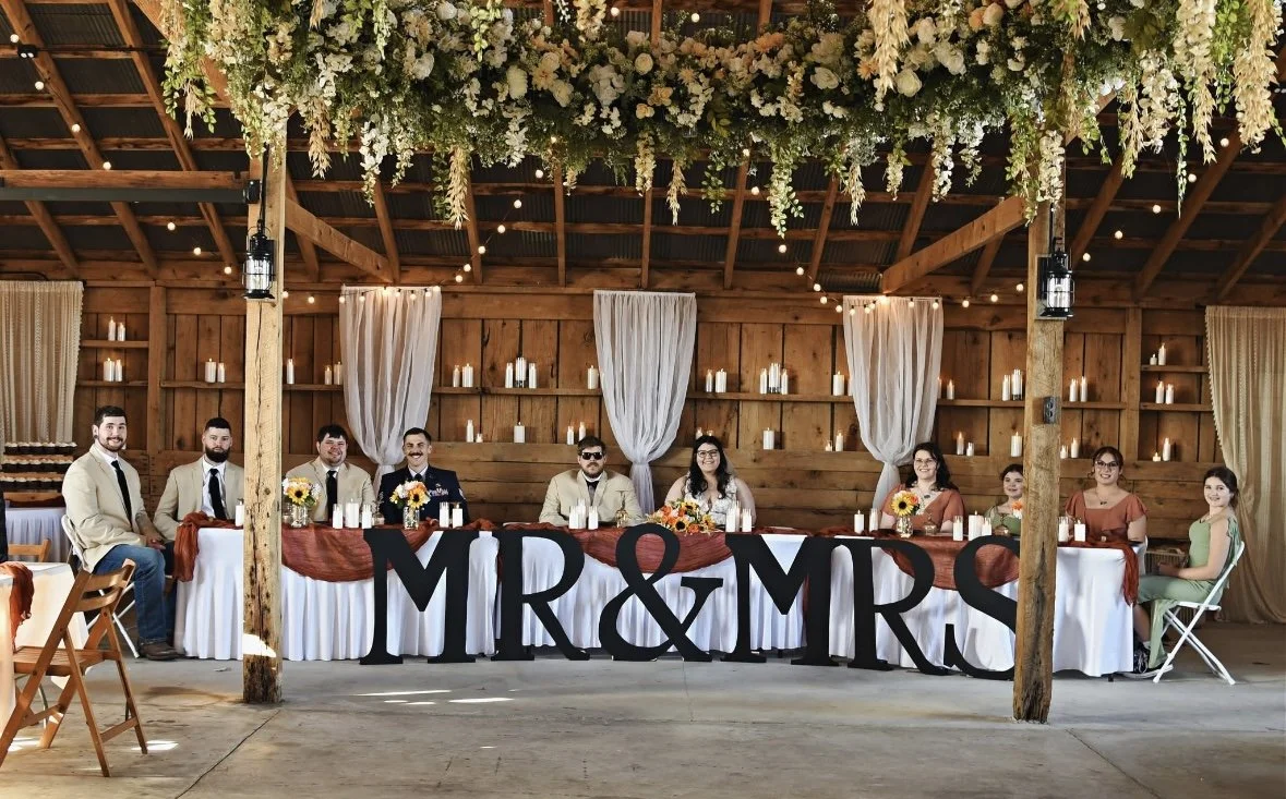 Wedding reception table with eight people seated behind it, decorated with candles, flowers, and a large sign that reads 'MR & MRS.' under a floral and wooden decor ceiling.