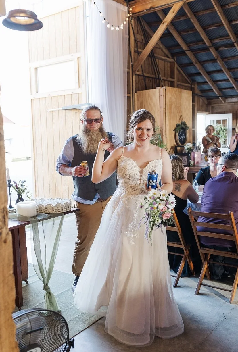 A bride in a white wedding gown holding a floral bouquet and a Bud Light can, smiling at a gathering in a rustic barn with wooden walls and ceiling, string lights, and guests sitting at a table.