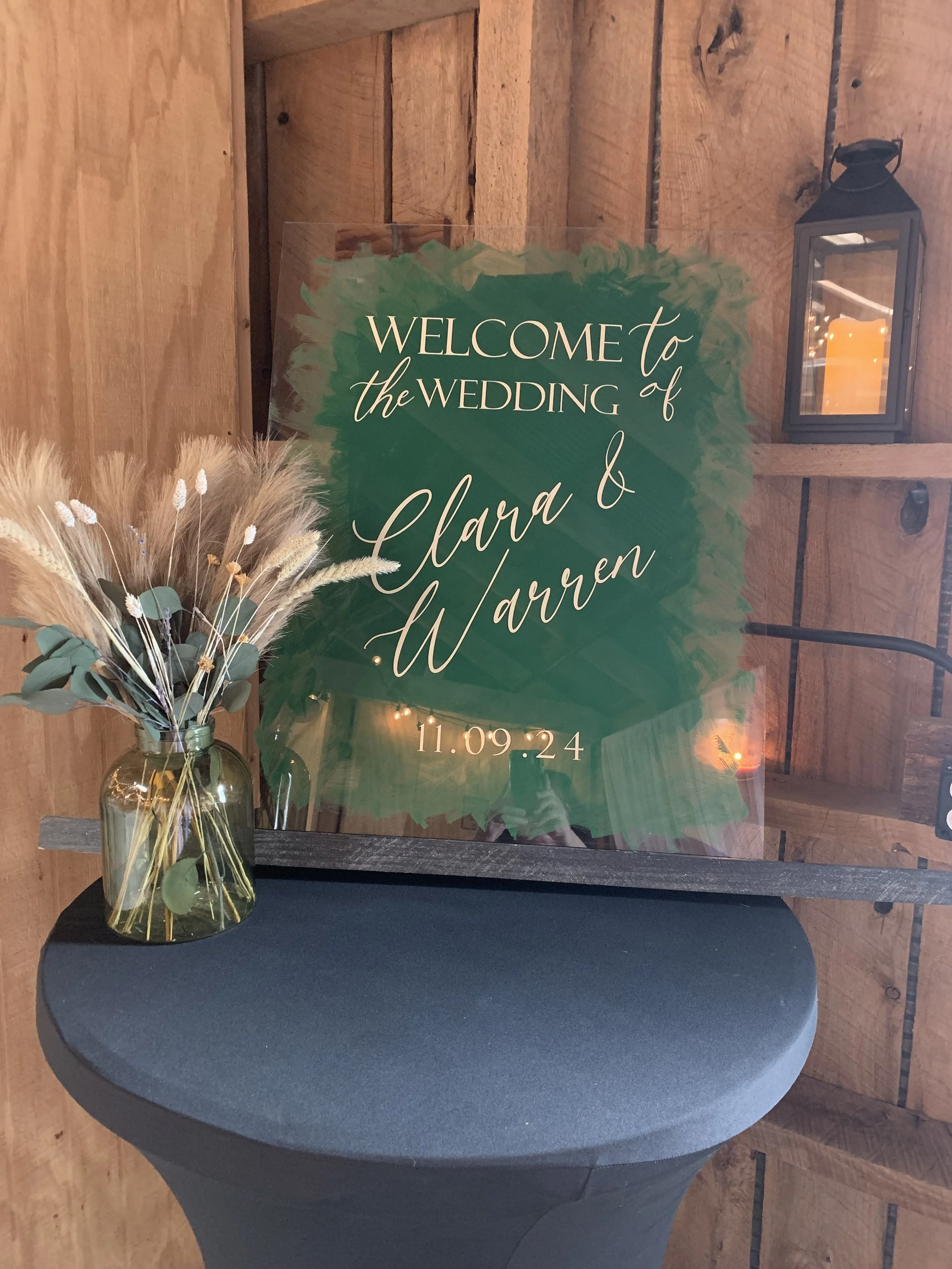 A welcome sign for a wedding on a clear acrylic board with green brush stroke background and white cursive text, reading 'Welcome to the wedding of Clara & Warren, 11.09.24'. There is a vase with dried flowers on a black table and a lantern on the wo