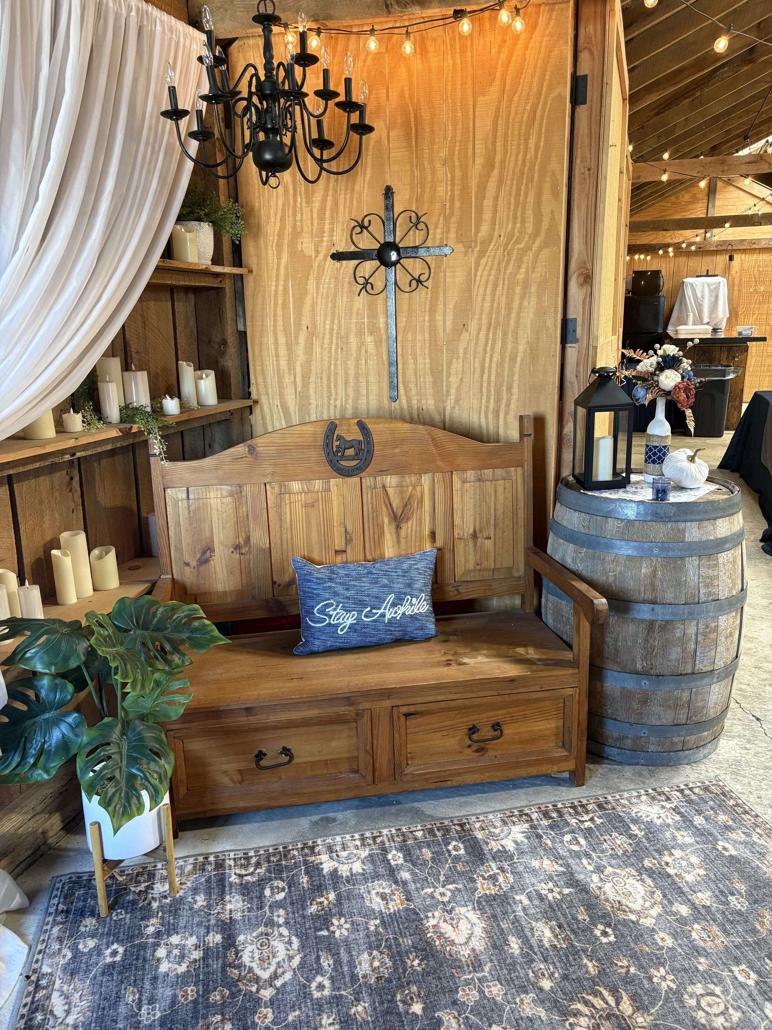 Rustic indoor decor with a wooden bench, a cushion saying 'Stay Agile', a chandelier, a wall with candles and cross, a barrel used as a table with a lantern, flowers, and small pumpkin, and string lights hanging from the ceiling.