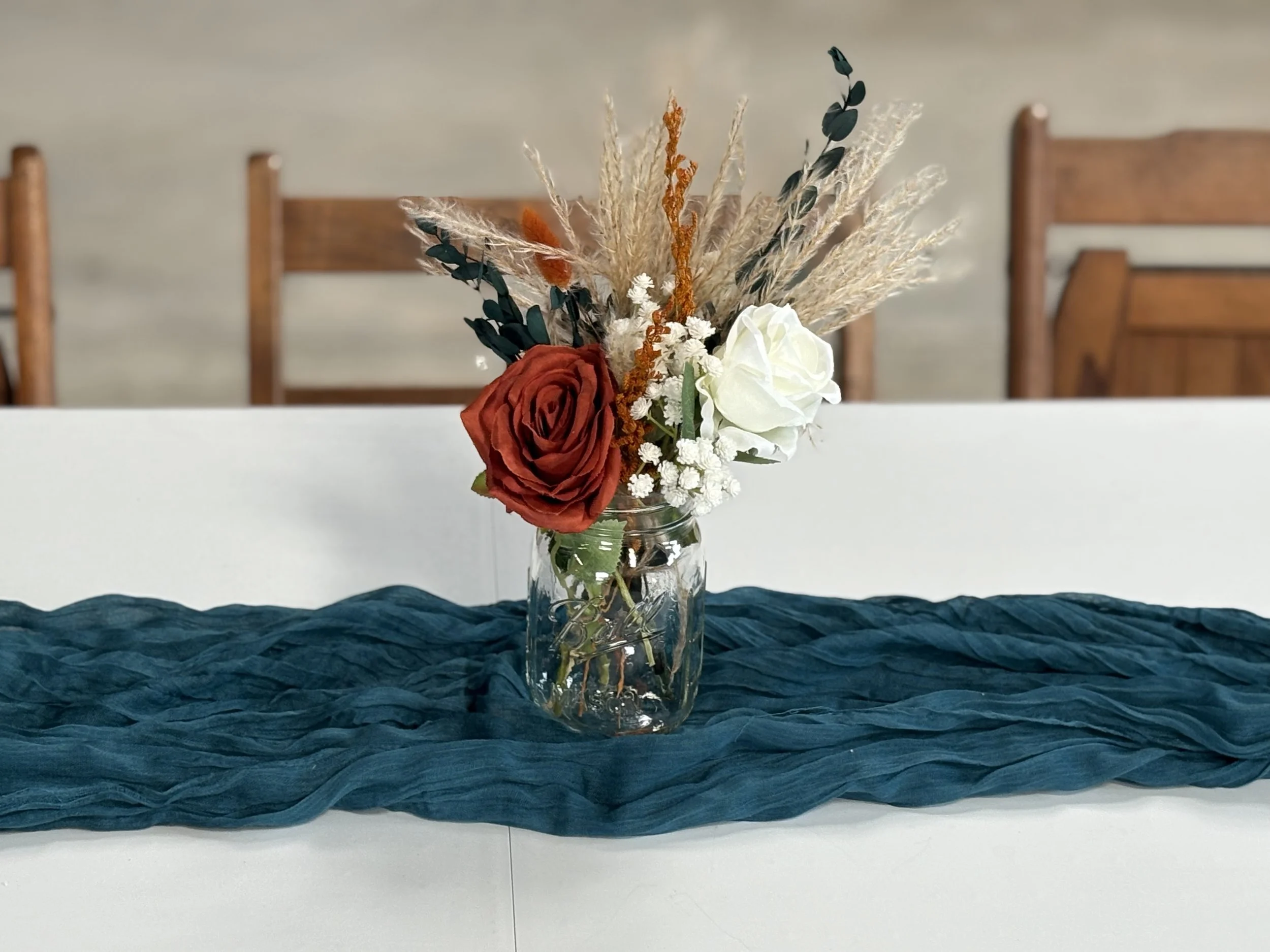 A glass jar with a bouquet of artificial flowers, including a red rose, white roses, and dried grasses, on a dark blue table runner on a white table, with wooden chairs in the background.