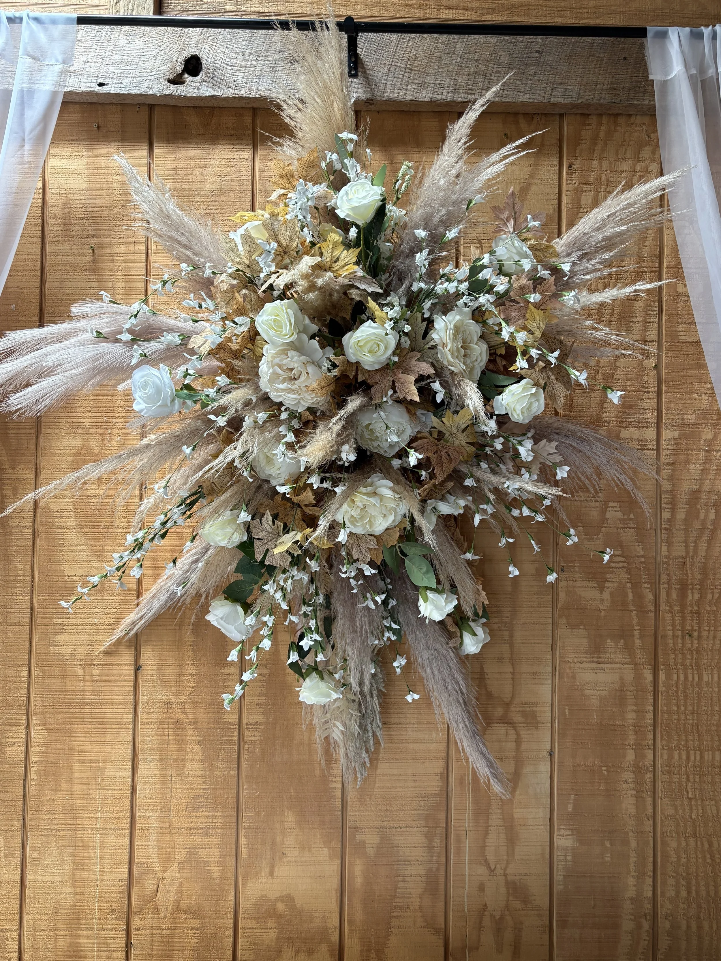 A floral arrangement with white roses, cream-colored petunias, gold and green leaves, and beige pampas grass, hanging on a wooden wall with white sheer curtains on either side.