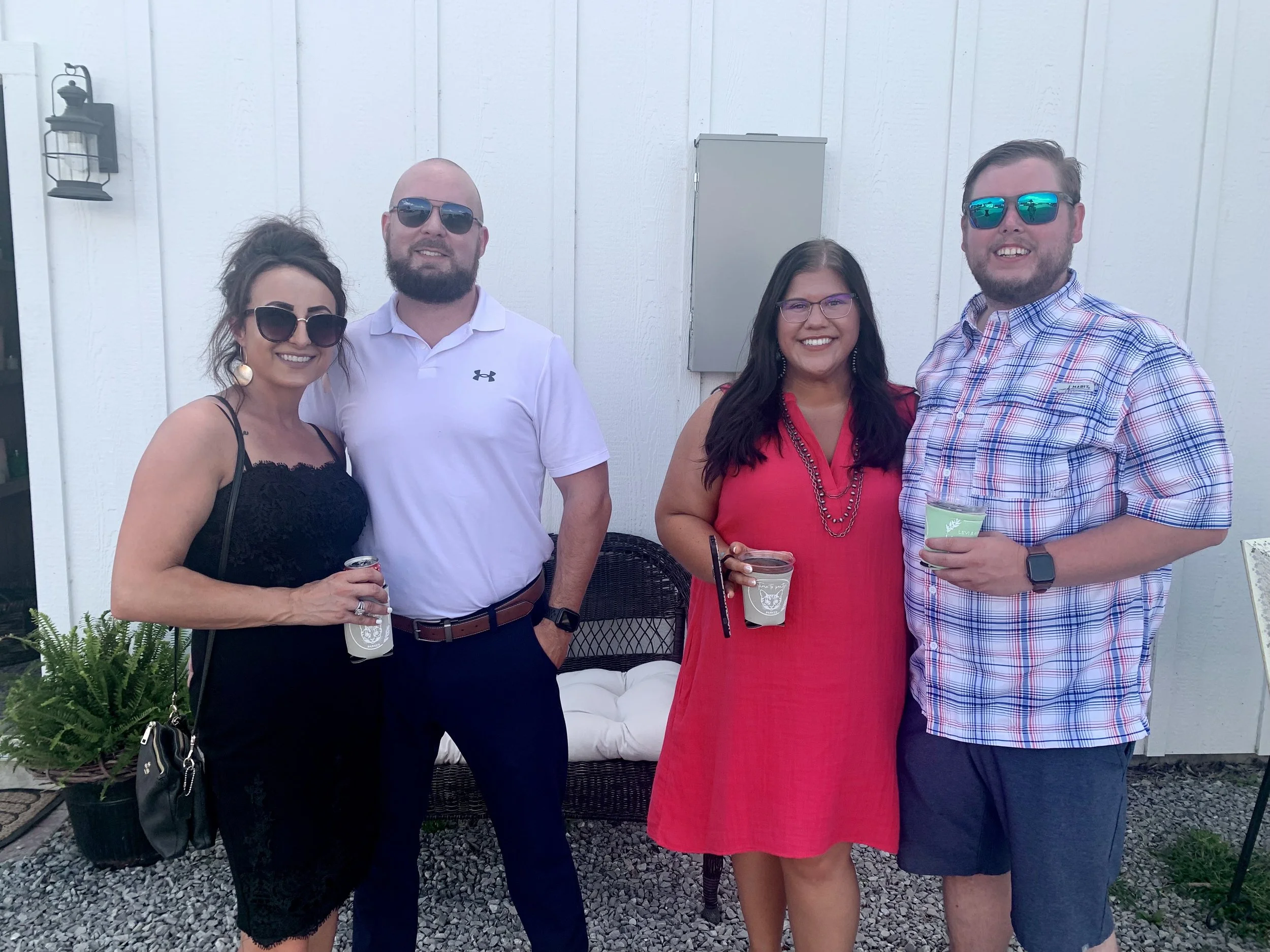 Four people standing outdoors in front of a white wall, smiling and holding drinks, enjoying a social gathering.