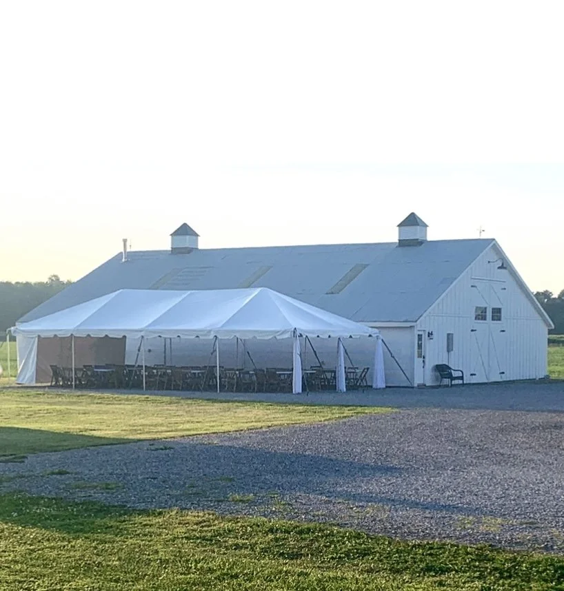 A large white barn with a tent set up next to it, multiple chairs, and tables inside the tent, set up outdoors on a grassy area.