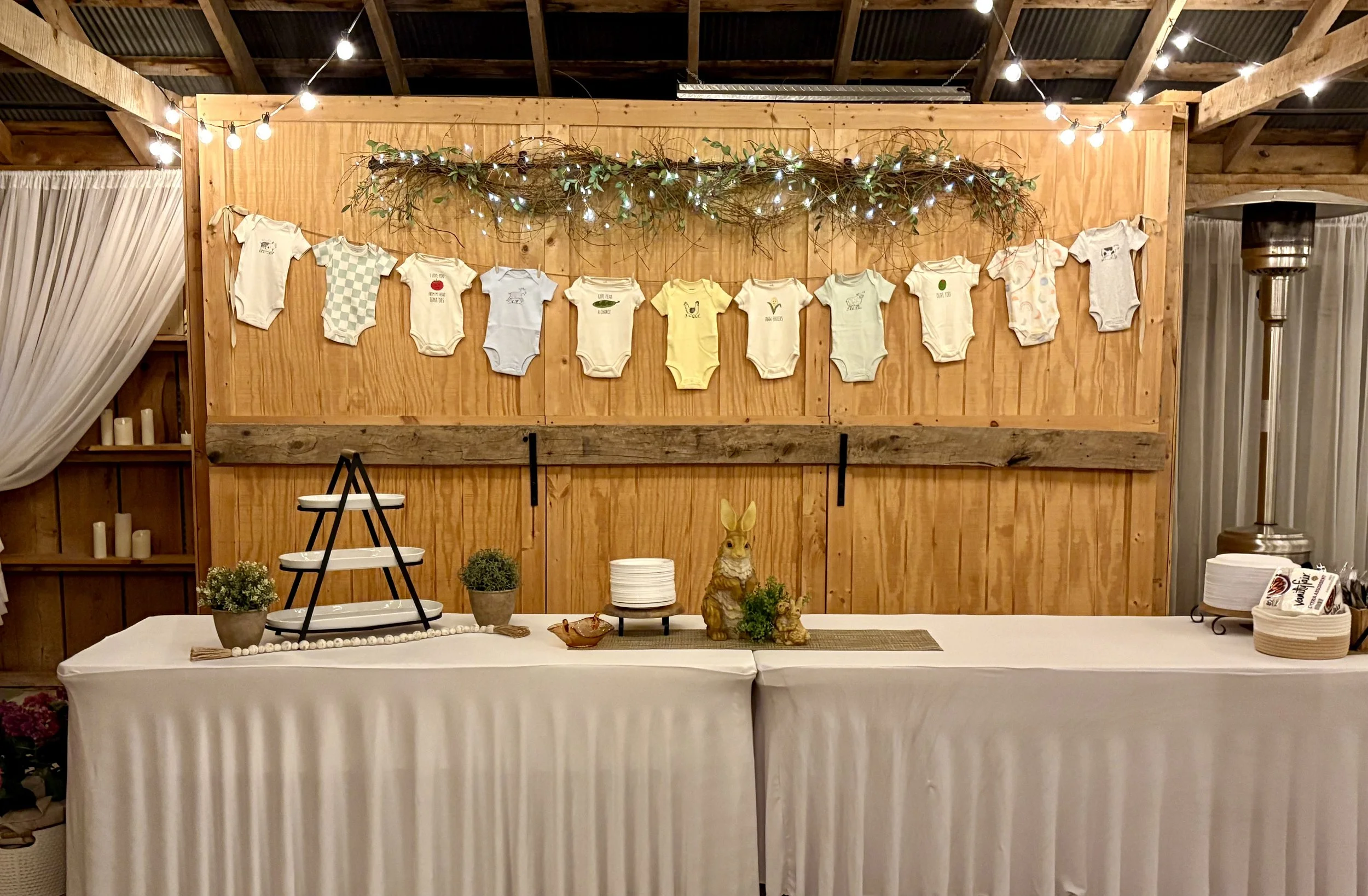 A dessert table at a baby shower with a white tablecloth, bunny figurines, small potted plants, plates, and a tiered stand. A wooden wall decorated with hanging baby onesies and string lights is in the background.