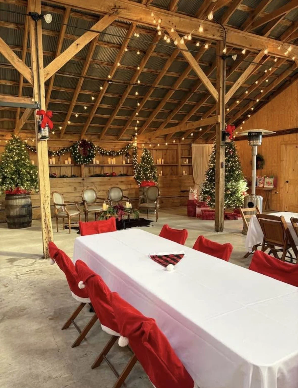 Decorated barn with Christmas trees, wreaths, string lights, and wrapped presents, set up for holiday celebration with tables and chairs.