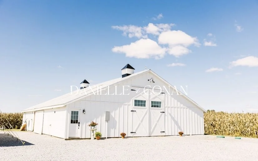 White barn with large doors and small windows, set in a gravel lot with a crop field in the background and a partly cloudy blue sky.