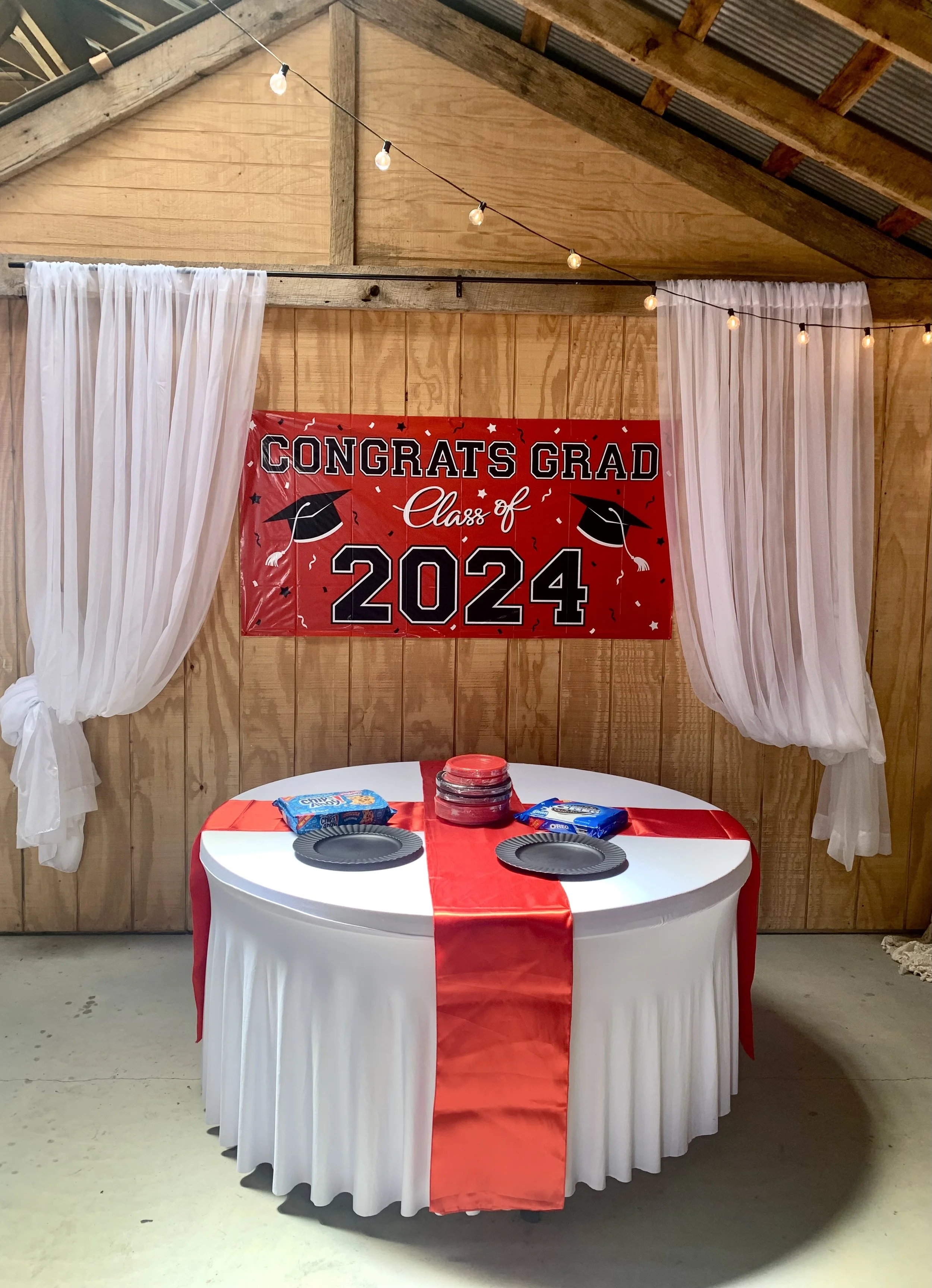 Graduation party setup with a round table covered in a white tablecloth and red runner, black paper plates, oreo cookies, red plastic cups, a red chipped plate, white curtains on either side, a wooden wall background, and a large red banner that read