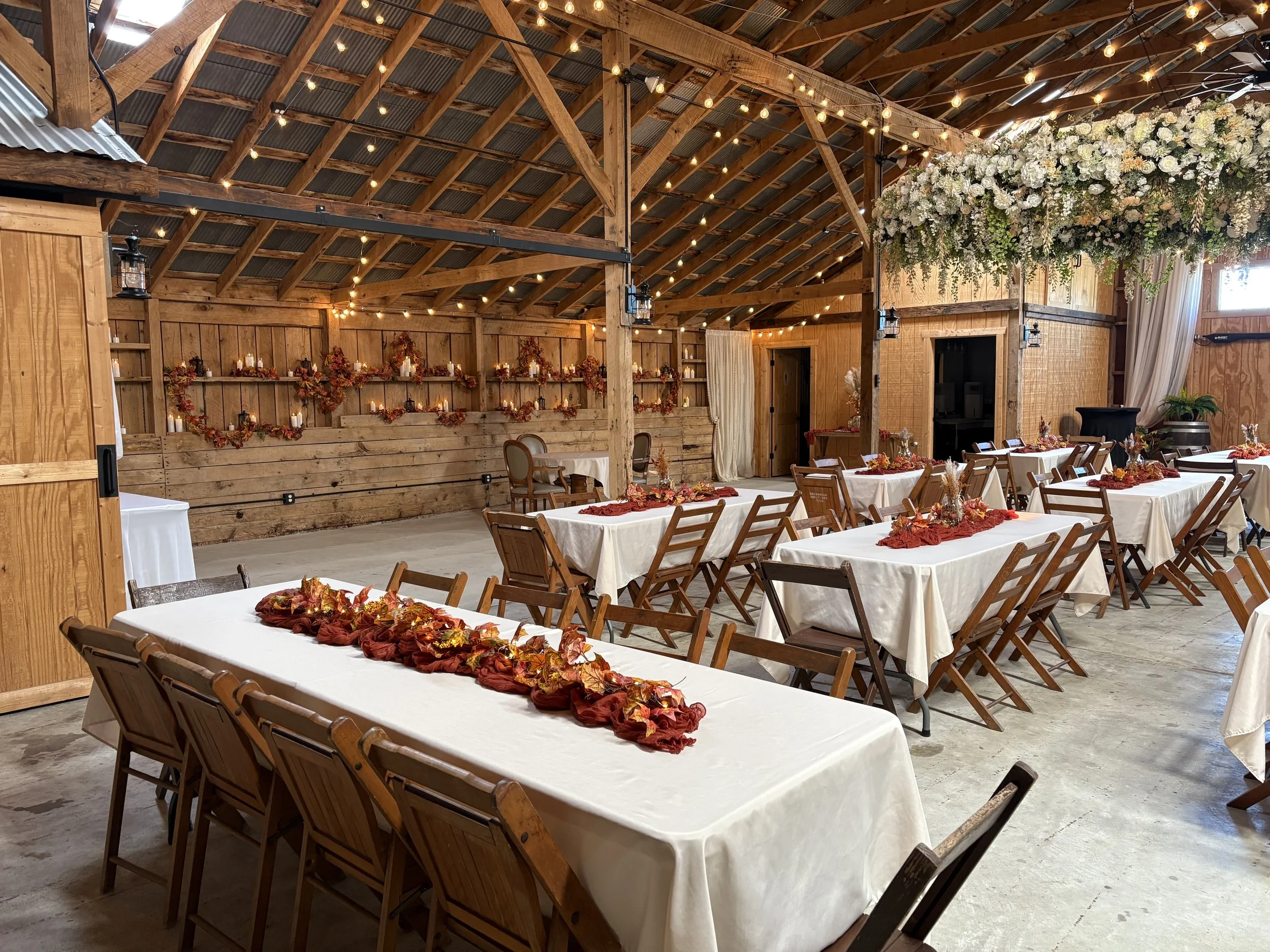 Wedding reception area with wooden decor, string lights, floral arrangements, and tables decorated with fall-themed centerpieces.