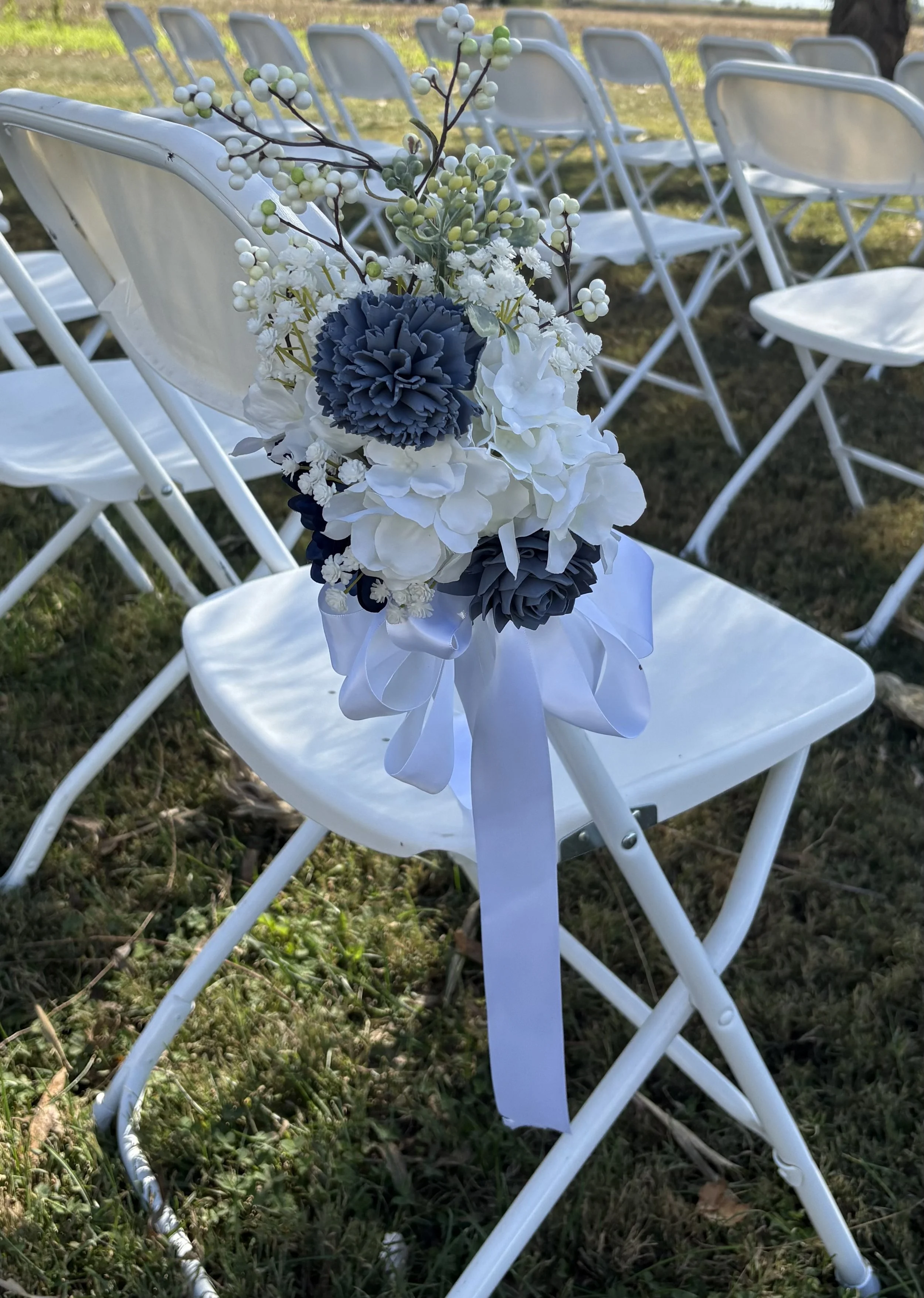 A white folding chair decorated with a flower arrangement attached to its side, consisting of white and gray artificial flowers and greenery, with white ribbon.