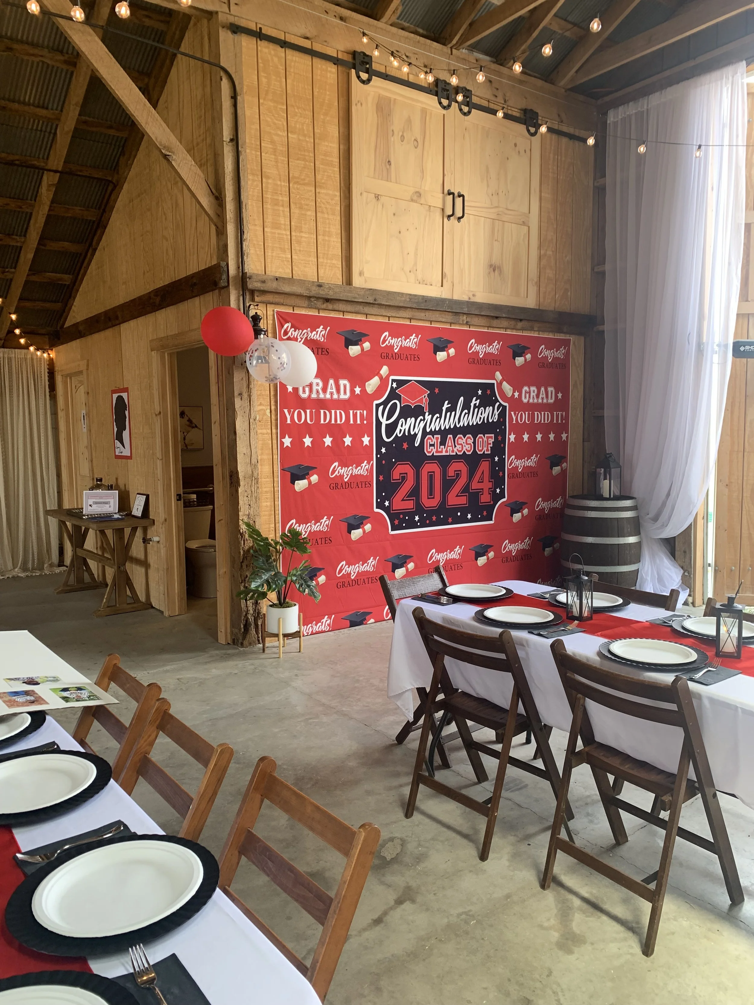 A decorated indoor space with a red and black graduation-themed backdrop that reads 'Congratulations Class of 2024' in a barn-style setting. There are tables with white tablecloths and black and white plates, lanterns, and chairs arranged for a celeb