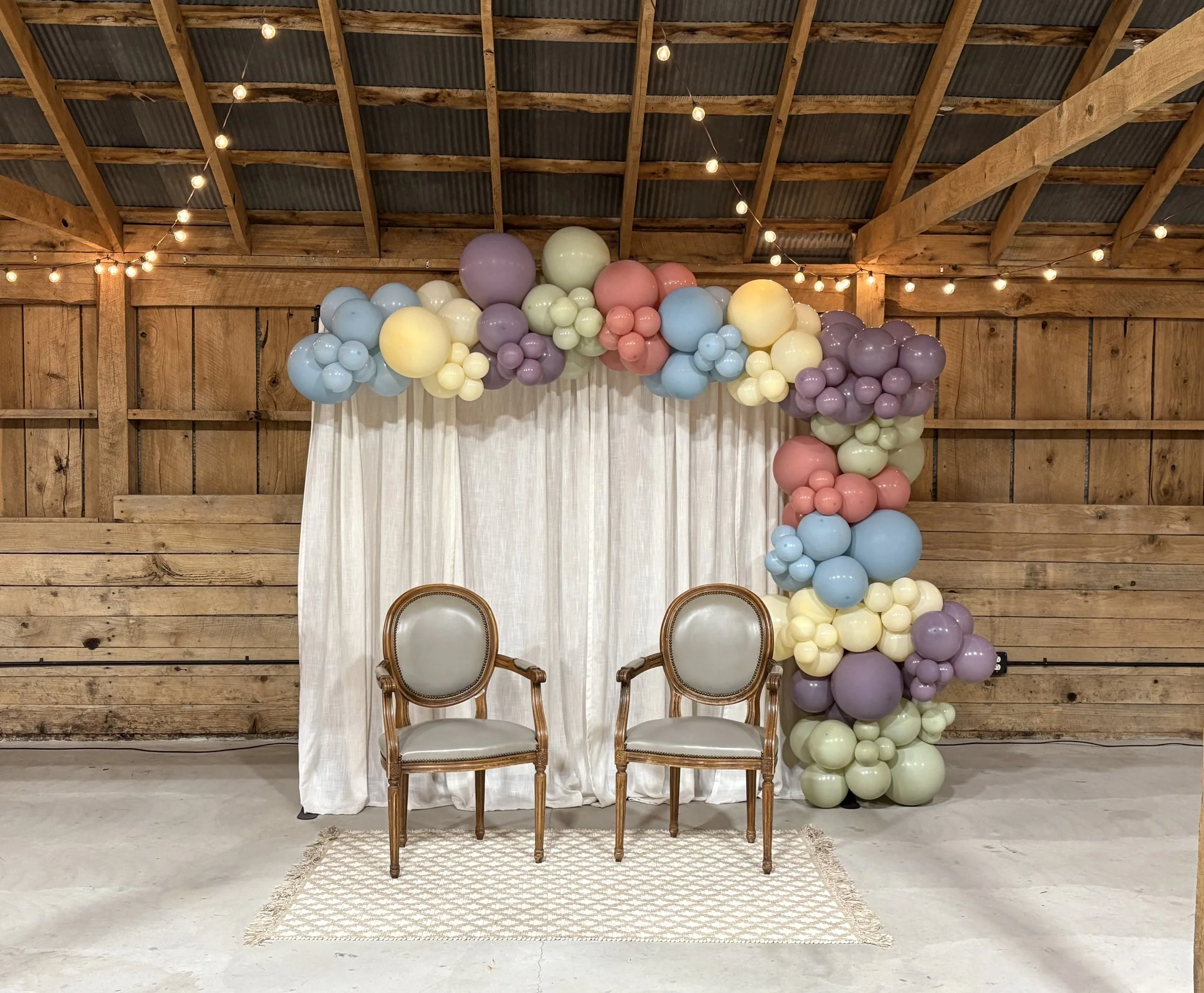Decorative setup with two vintage chairs in front of a white curtain, adorned with pastel-colored balloons arranged in an arch and cluster, with string lights hanging from a wooden ceiling in a rustic wooden barn setting.