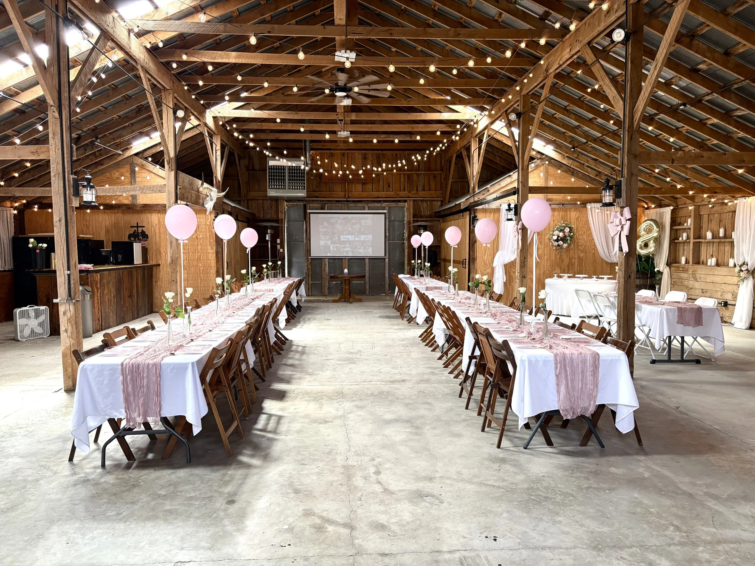 An event space in a rustic barn with wooden beams and string lights, decorated with pink balloons and white flowers, set up with long banquet tables for a celebration.