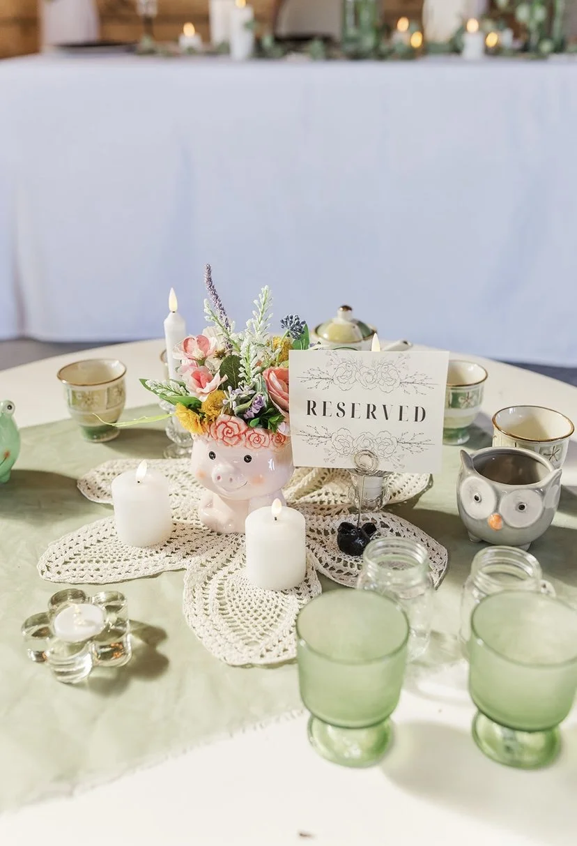 Table setting with a floral centerpiece in a pig-shaped vase, surrounded by candles, cups, and glasses, with a reserved sign.