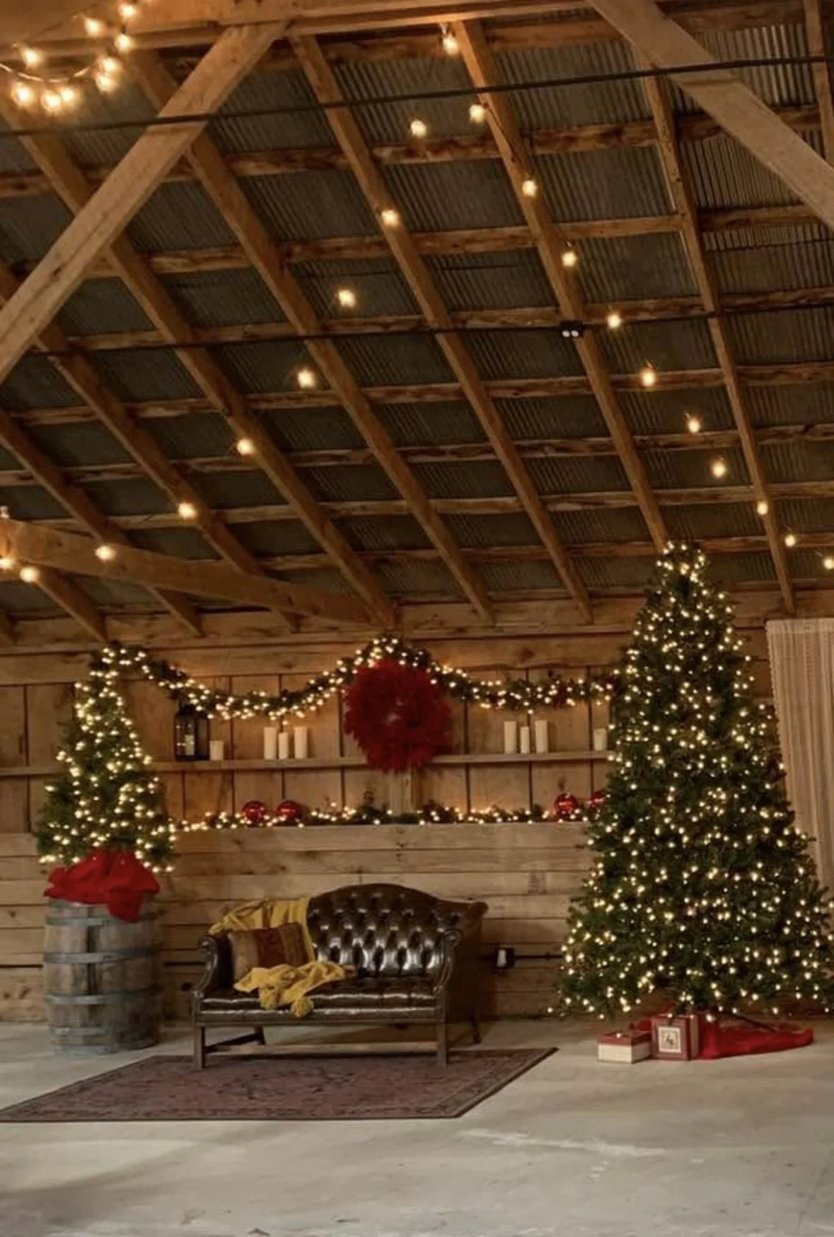 Decorated Christmas tree with presents underneath, two small Christmas trees, and festive garland with lights in a rustic wooden barn setting.