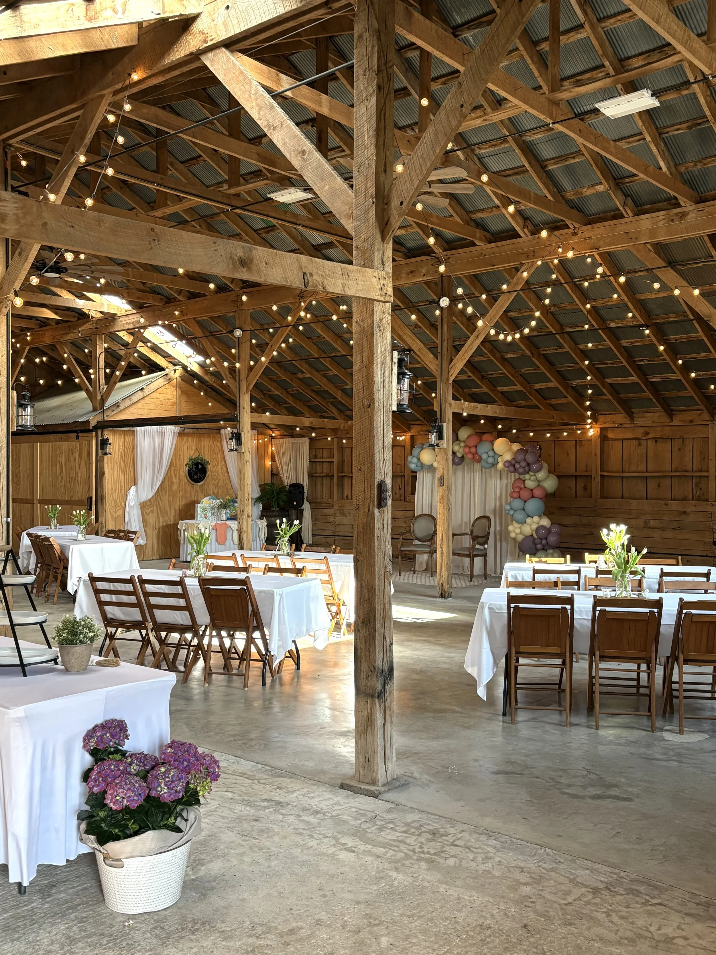 Wedding reception setup in a rustic barn with white tablecloths, flower arrangements, string lights, and a balloon arch.