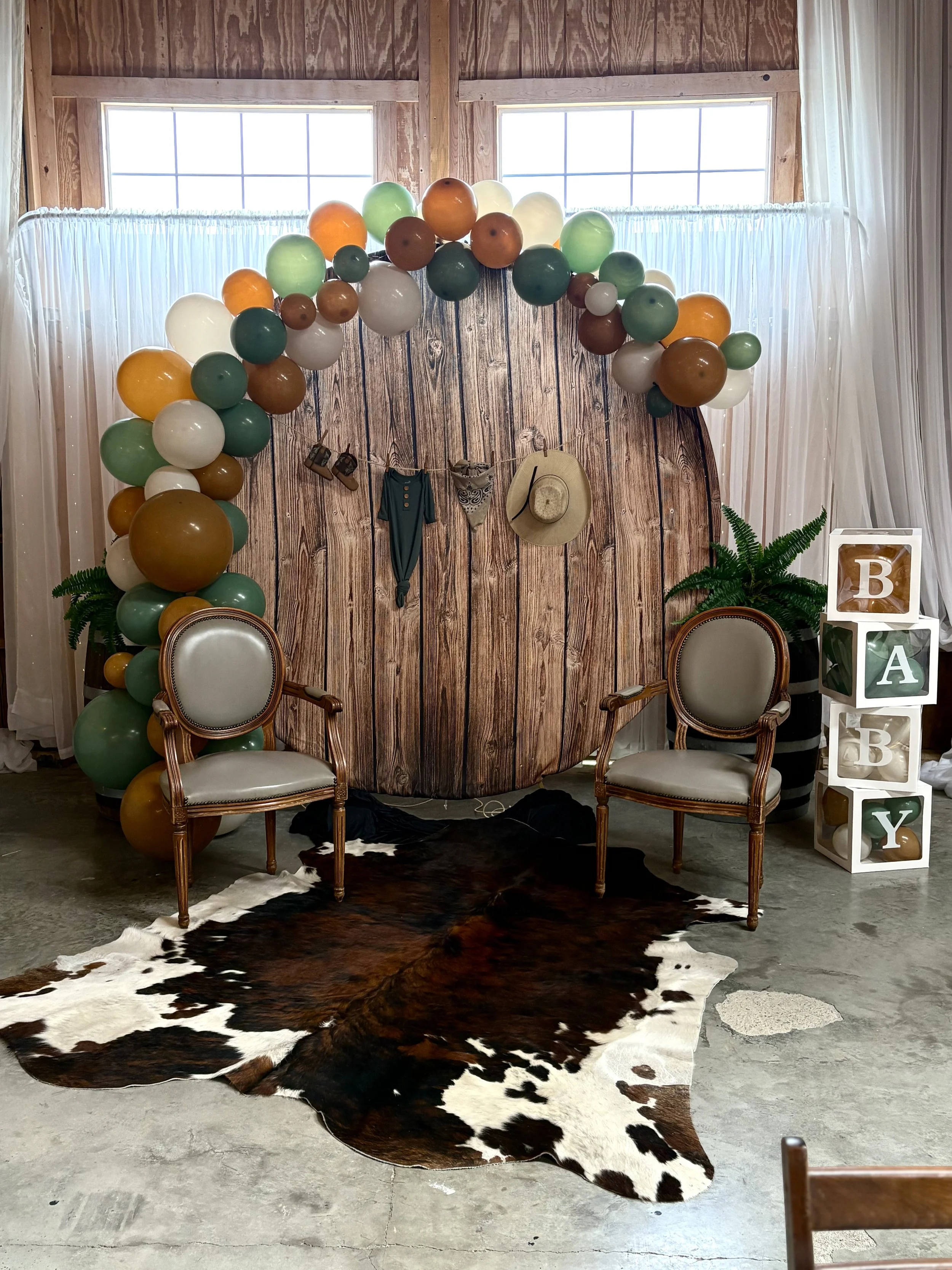 A rustic wooden backdrop decorated with multicolored balloons in earth tones, two vintage armchairs, a cowhide rug on a concrete floor, and a white cube storage unit with blocks spelling 'BABY' on the right, with small plants on either side of the ba