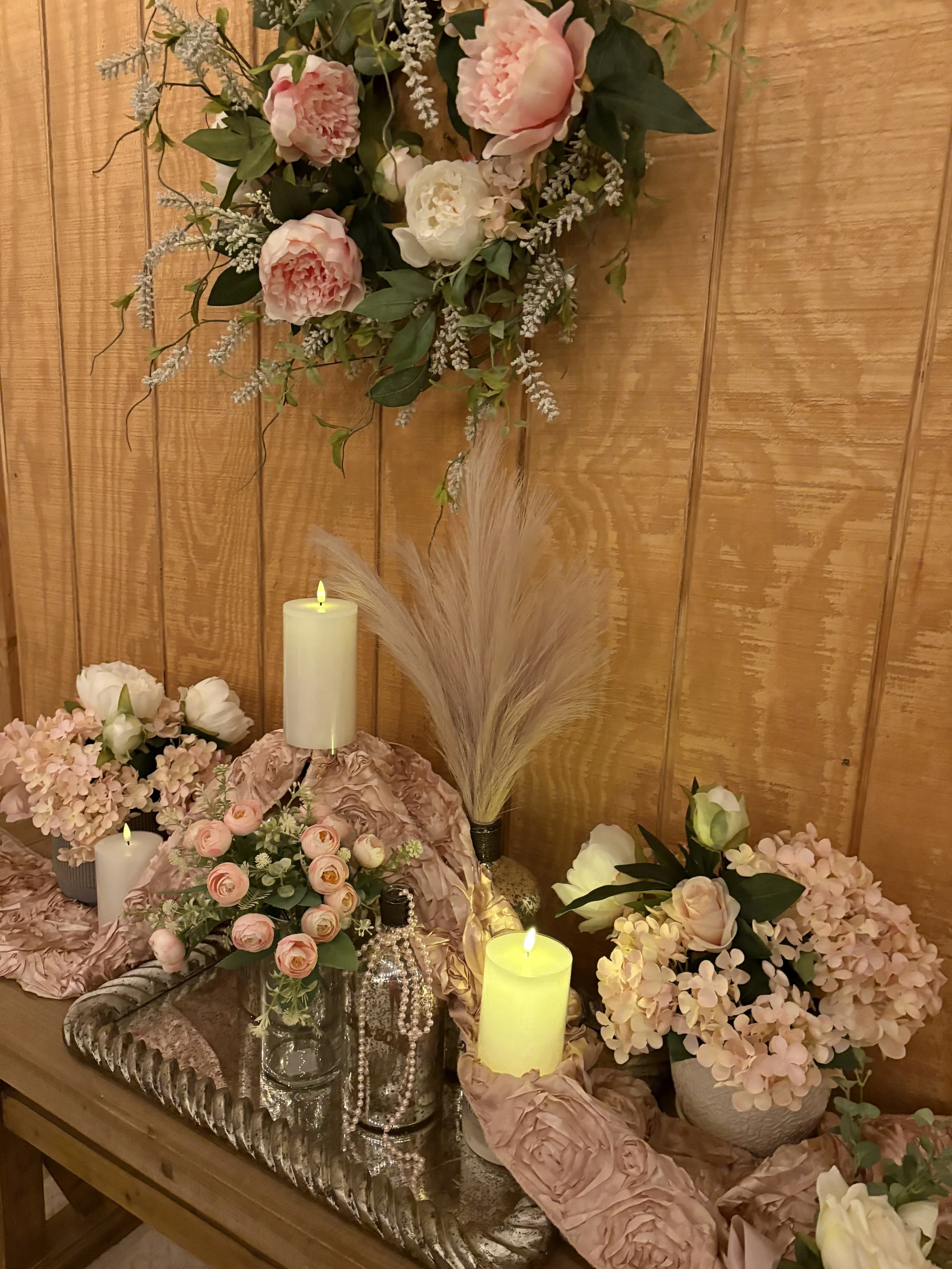 Arrangement of pink and white flowers, candles, and decorative bottles on a wooden table against a wooden wall.