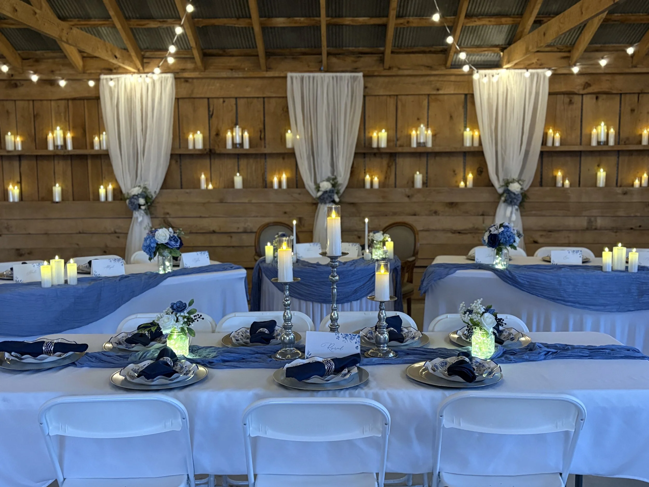 Elegant wedding reception setup with long tables covered in white tablecloths, blue runners, floral centerpieces with white and blue flowers, and candles. The background features a wooden wall with white curtains and numerous small candles on shelves