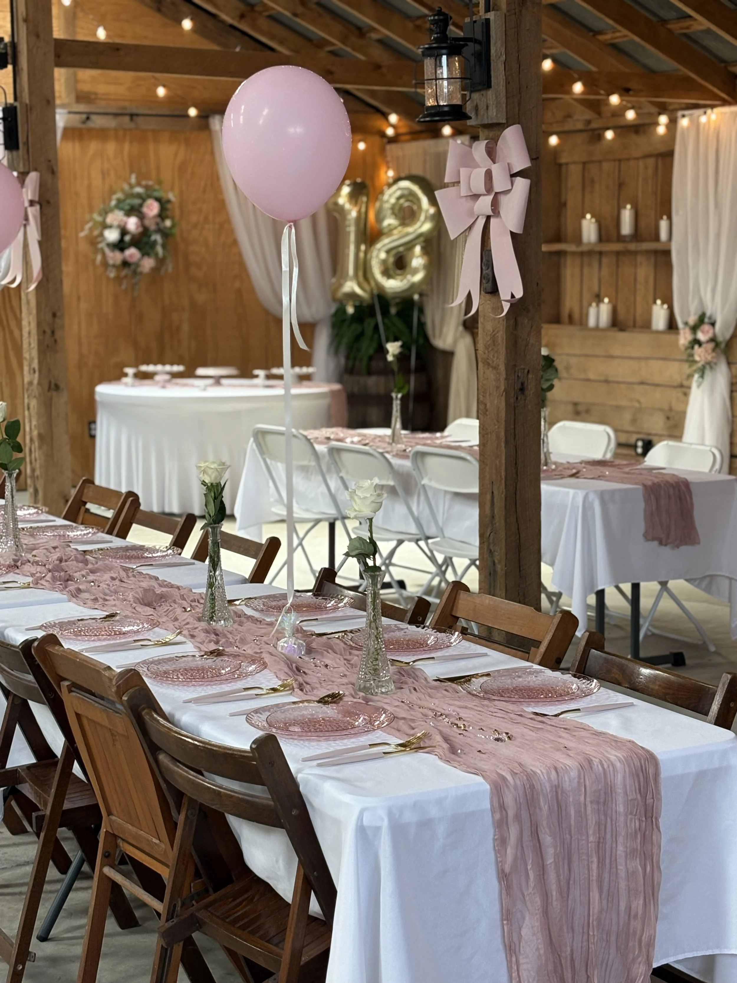 Decorated indoor wedding reception with a long table covered with a pink table runner, pink glass plates, gold utensils, white napkins, and white roses in glass vases. Pink balloons and a large gold '18' balloon in the background. Wooden walls and ceiling, with string lights and candles.