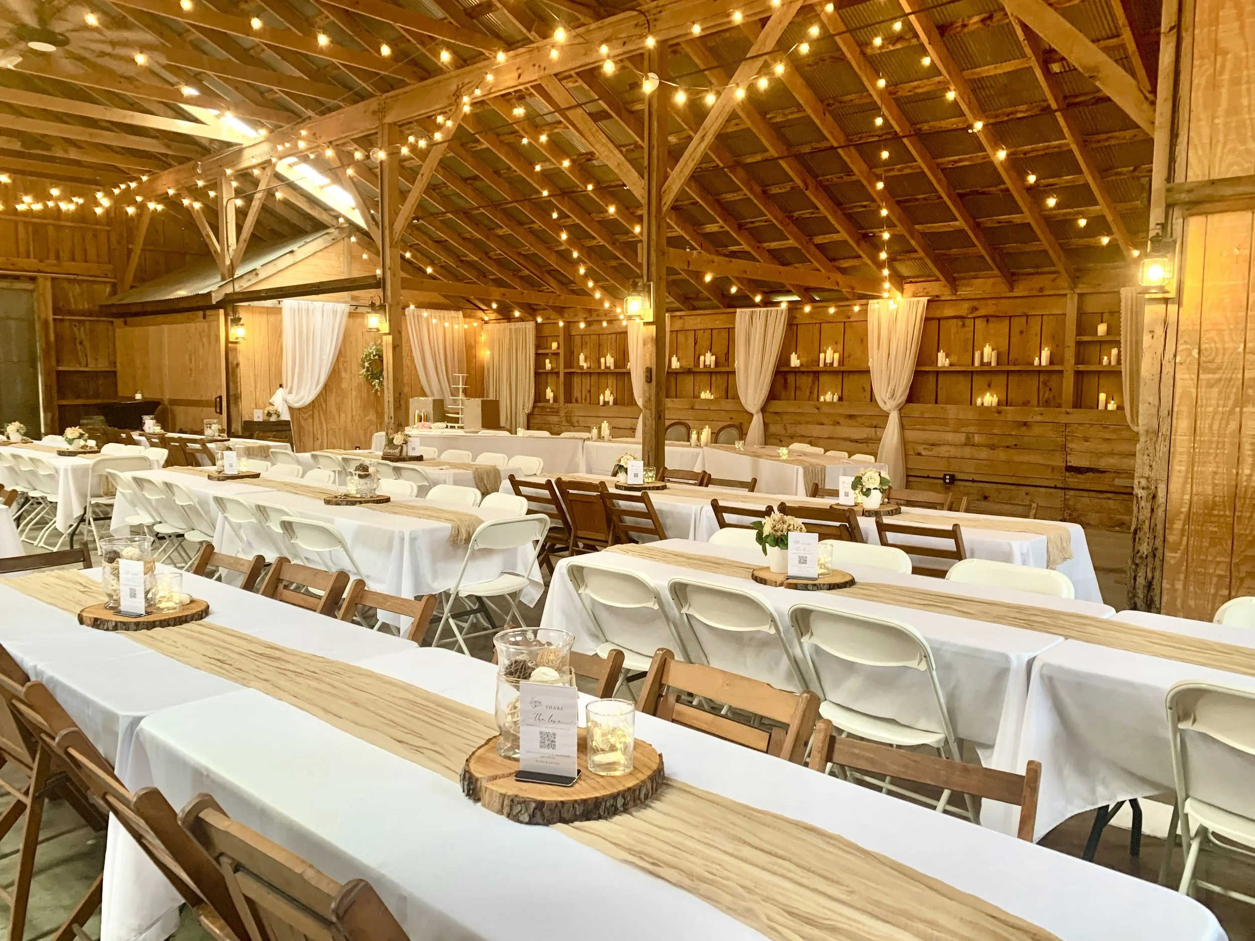 Inside a rustic barn decorated for a wedding reception with string lights, candles, and long tables with white tablecloths and wooden accents.