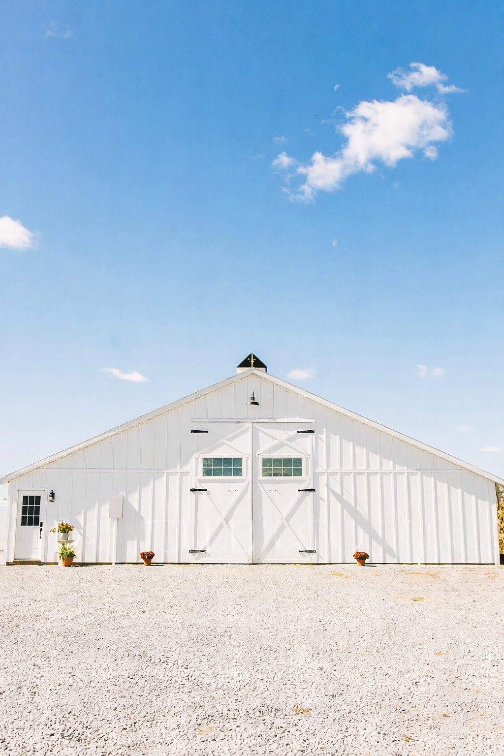 A white barn with large double doors, small windows, and potted plants outside under a blue sky with scattered clouds.