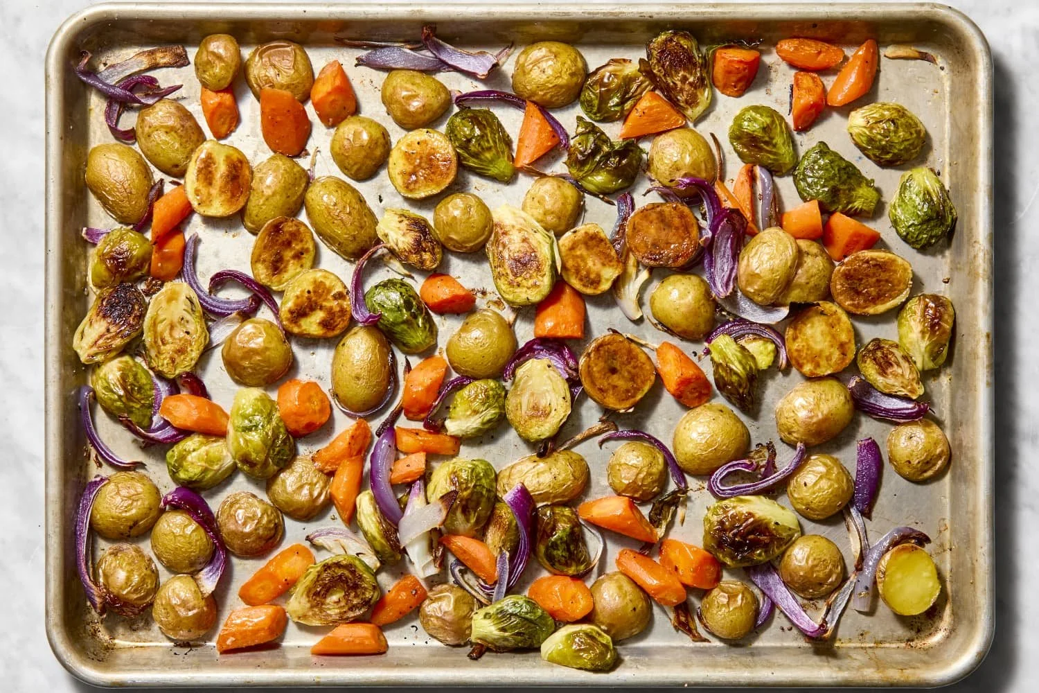 Roasted baby potatoes, chopped carrots, red onions, and Brussels sprouts on a baking sheet.