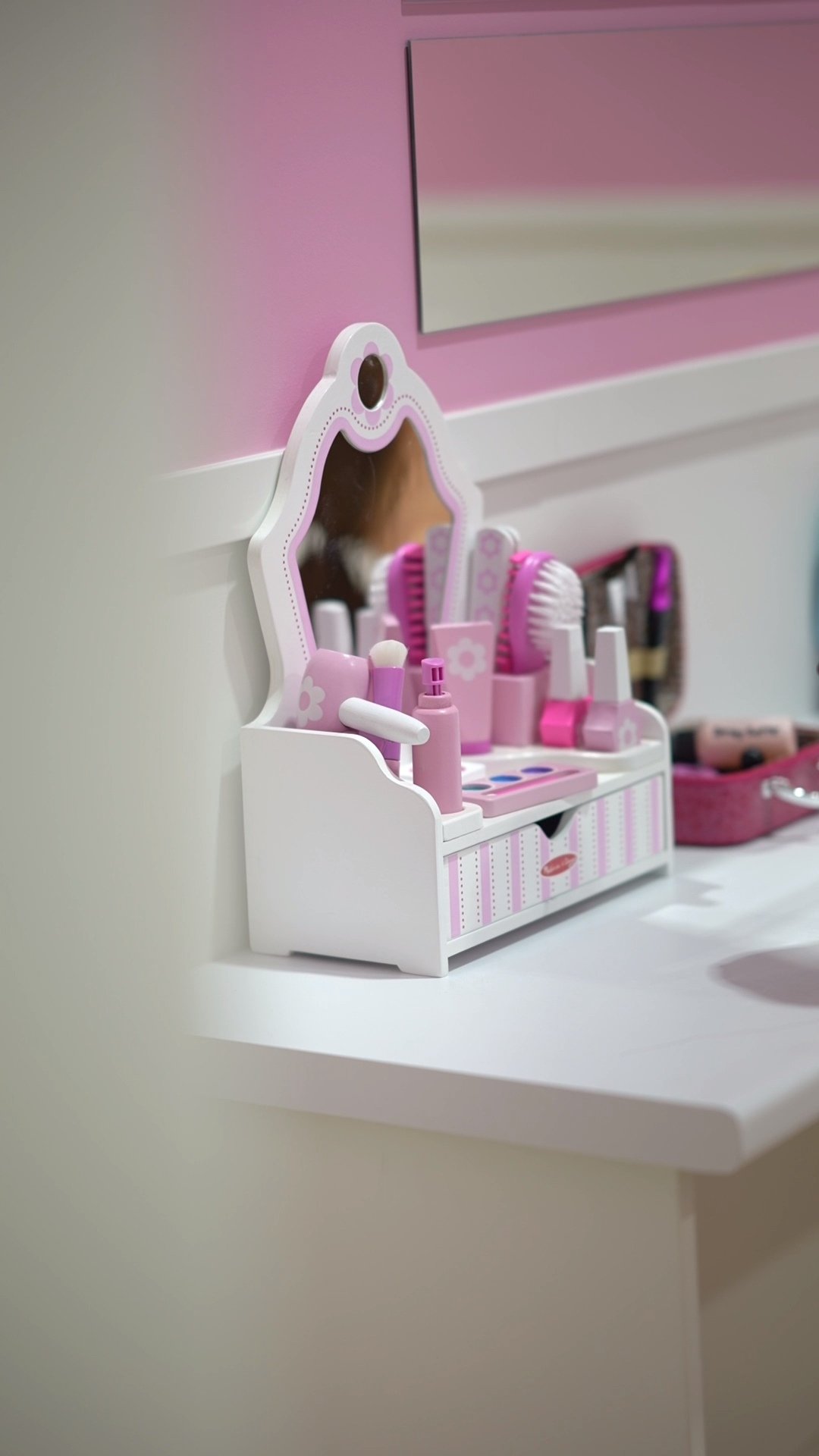 Pink and white toy vanity with mirror and miniature makeup items, on a white surface against a pink and beige wall.