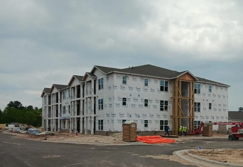 Three-story apartment building under construction, wrapped in Tyvek with scaffolding and construction equipment nearby. Workers are present on-site.