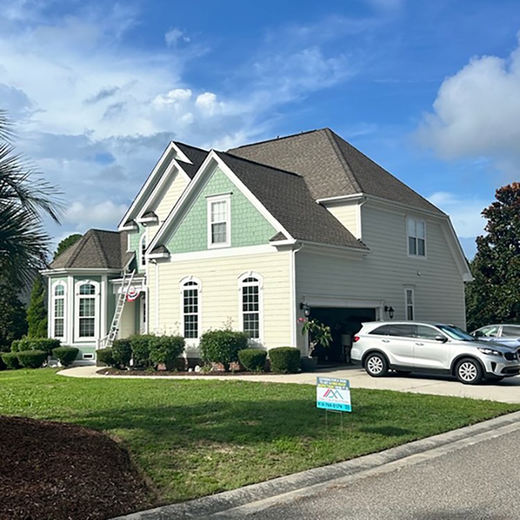 Suburban house with a two-car garage, ladder against the facade, lawn, and parked cars. Cloudy blue sky overhead with a sign in the yard.