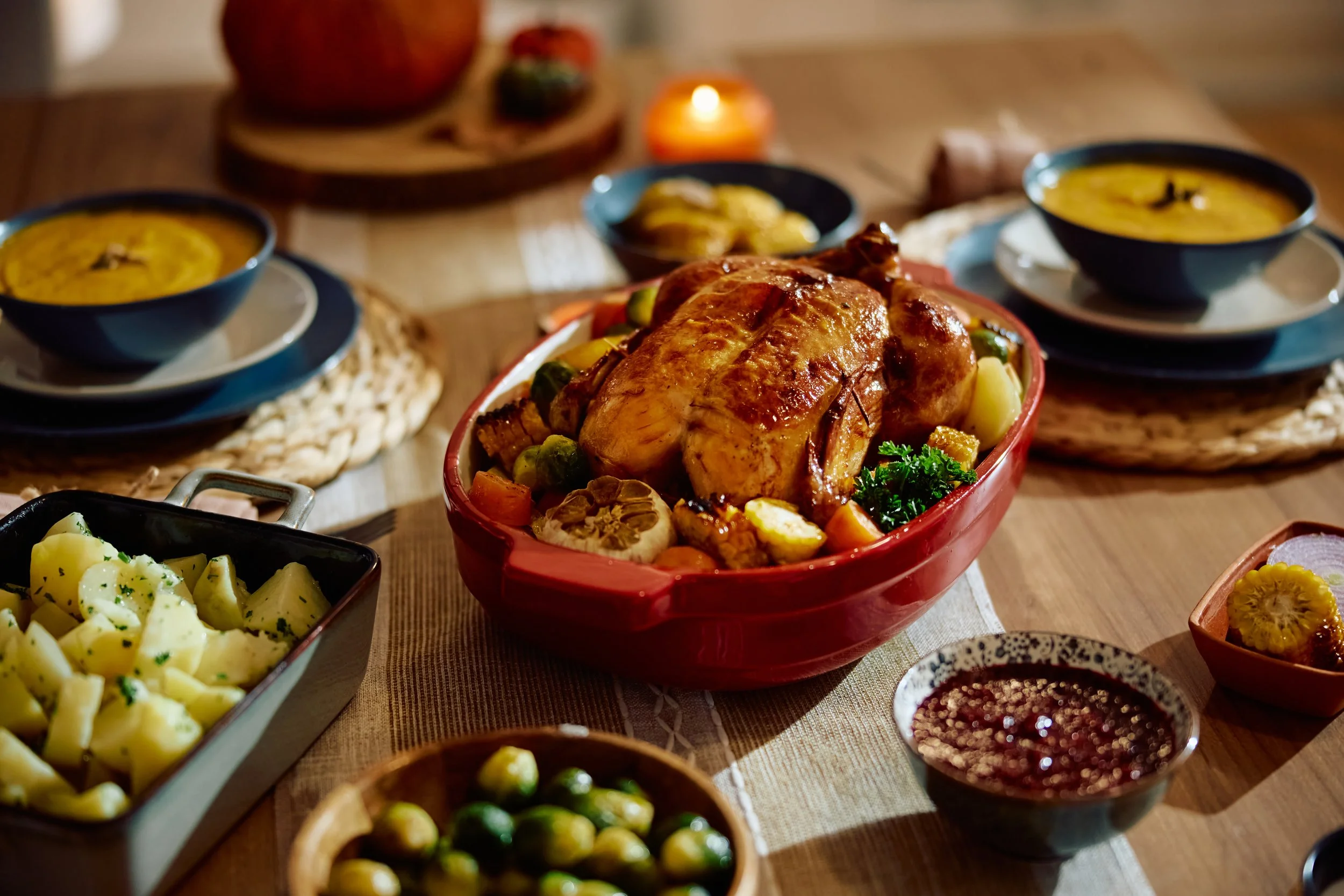 A festive dinner table with roasted chicken in a red dish surrounded by vegetables, bowls of soup, mashed potatoes, and condiments.