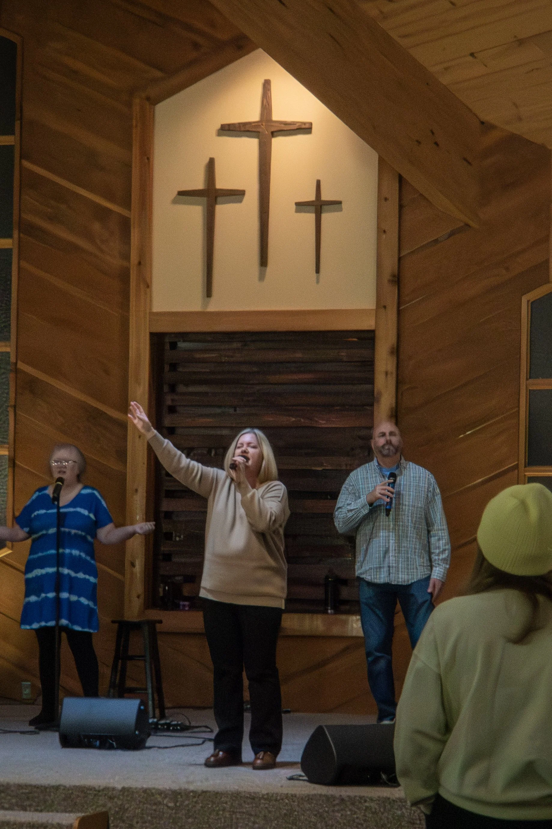 A church service or worship event with three people singing on stage, one woman holding a microphone with one arm raised, in a wooden interior with three wooden crosses on the wall behind them, and three congregation members in the foreground.