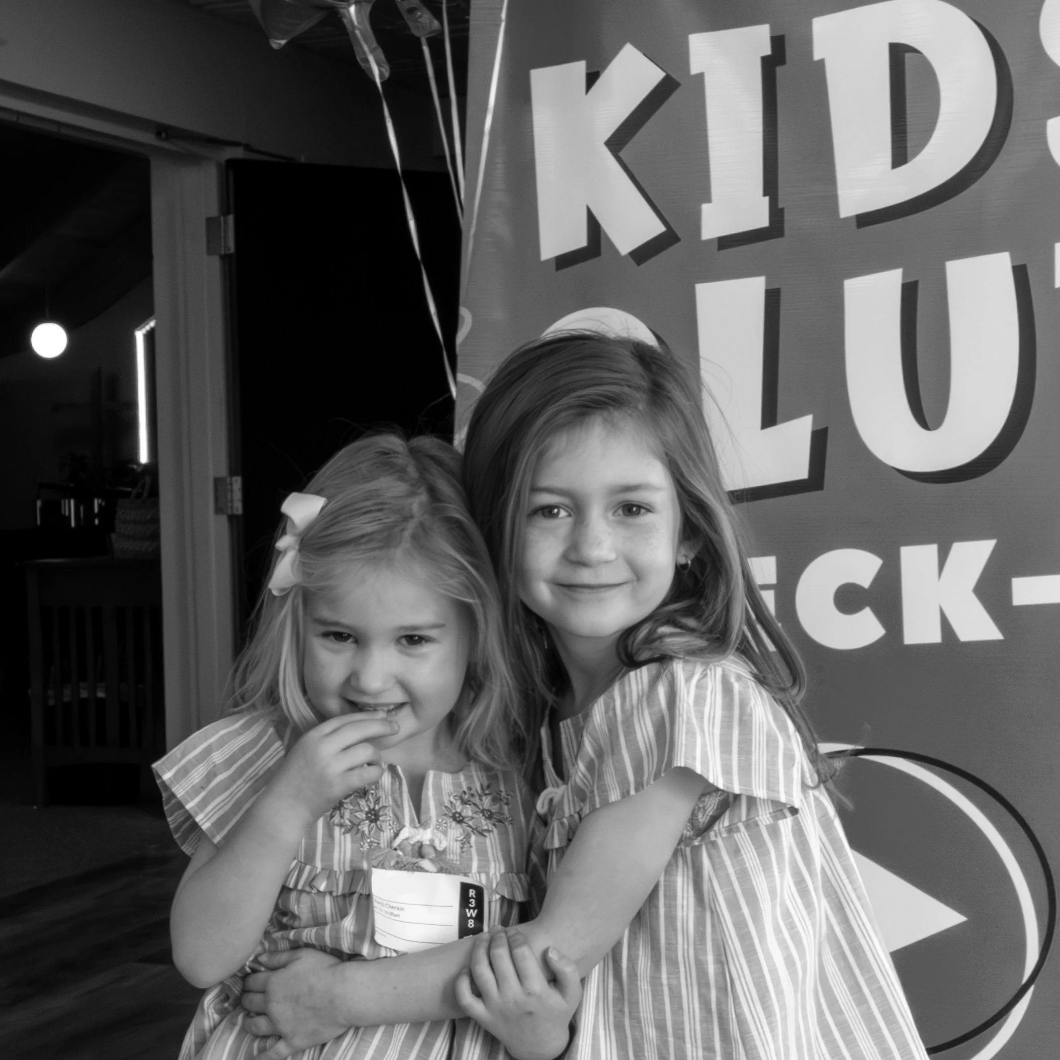 Two young girls hug each other, smiling at the camera at a 'Kids Lunch' event. The younger girl has a bow in her hair and is eating a snack.