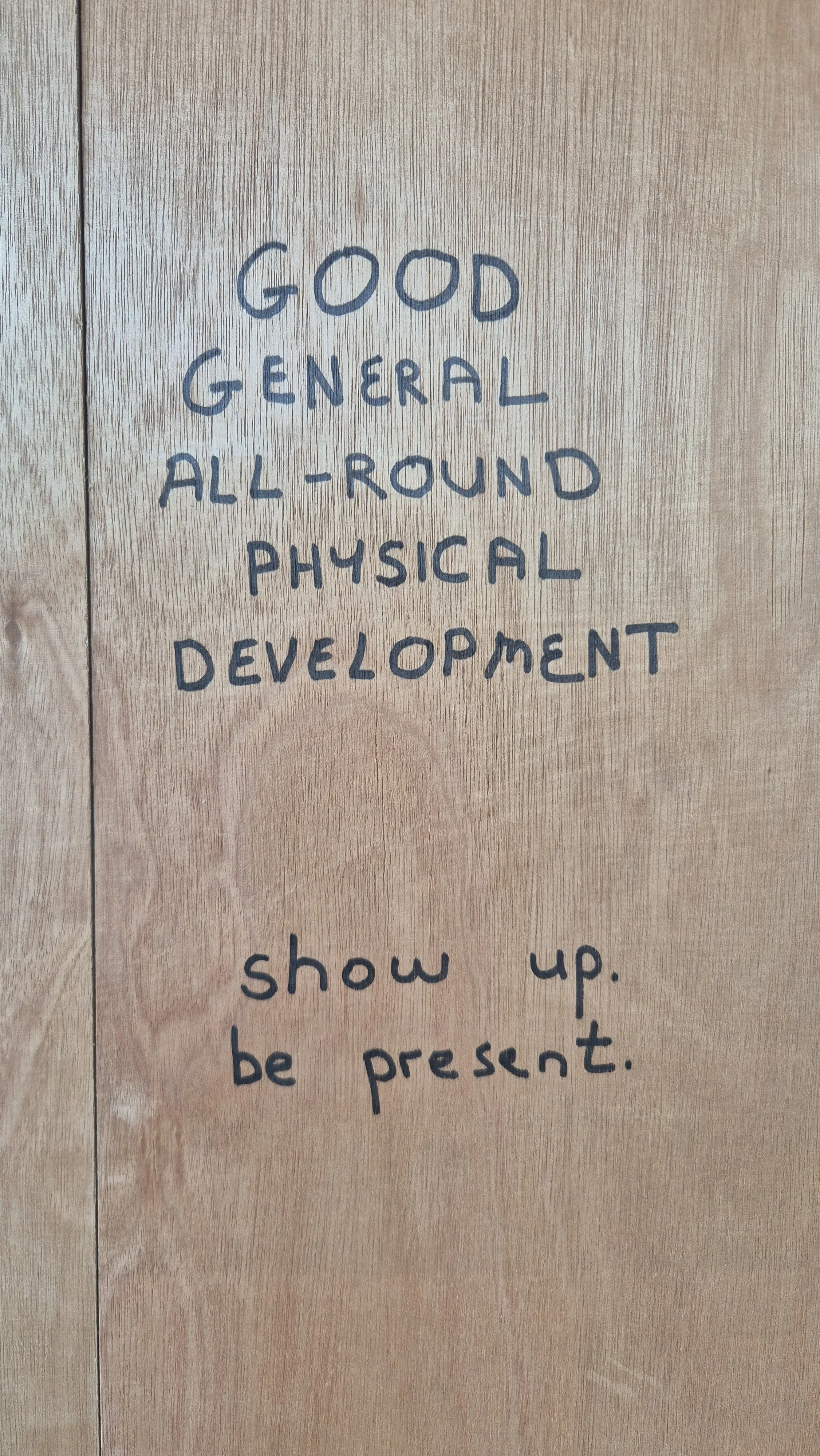 Handwritten sign on wood surface with the text: "Good general all-round physical development, show up, be present."